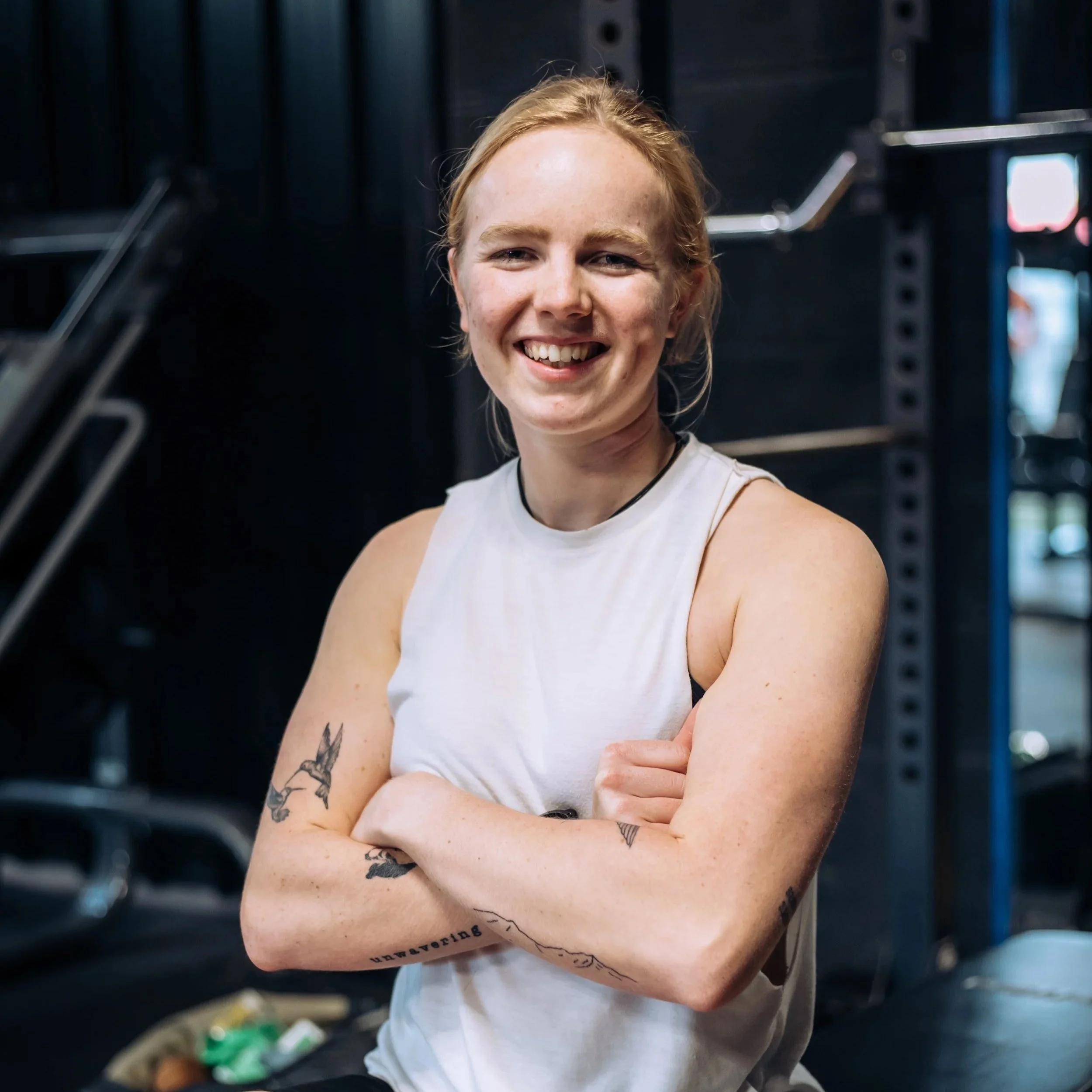 A young woman smiling with crossed arms, tattoos on her forearms, standing in a gym with dark equipment in the background.