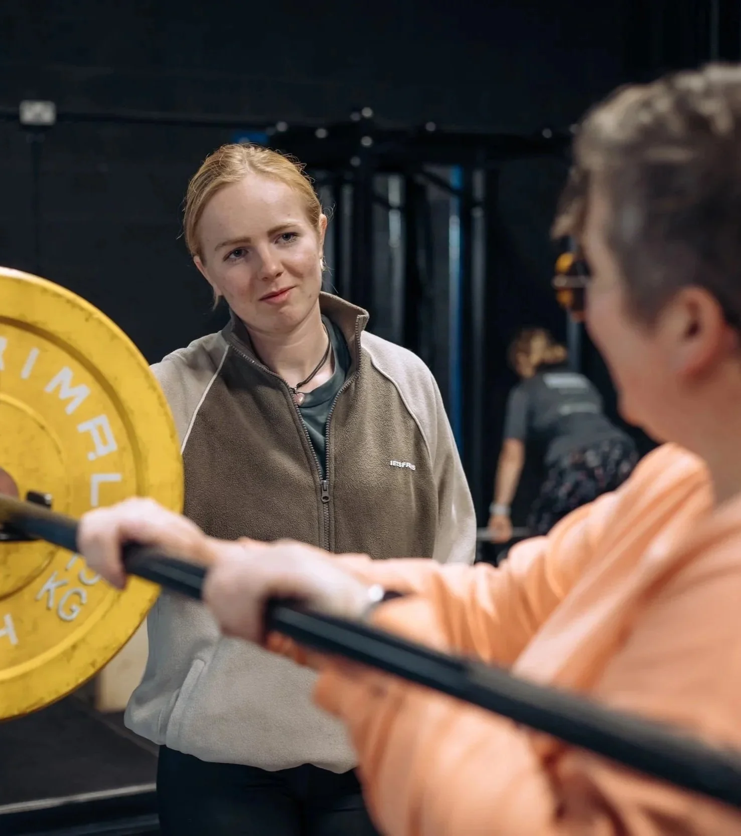 Two women lifting a barbell with a yellow weight plate in a gym, with one woman looking at the other.