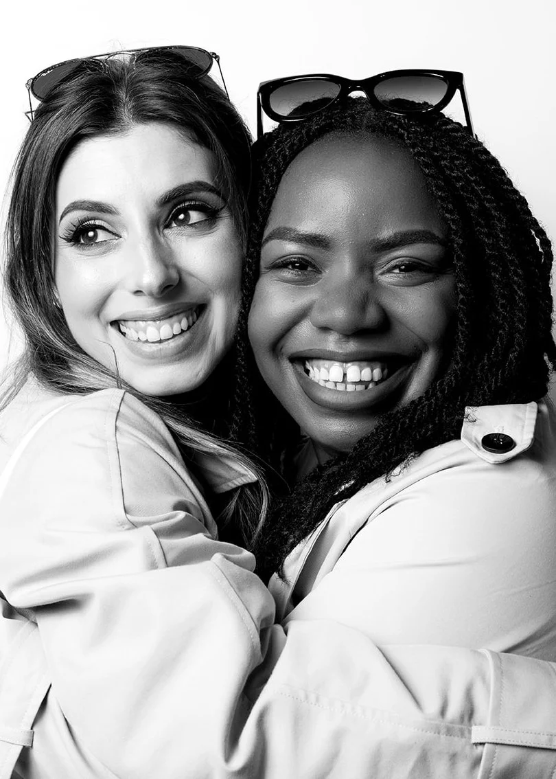 Two women smiling and hugging, both wearing light-colored jackets, with sunglasses resting on their heads, in a black-and-white portrait.