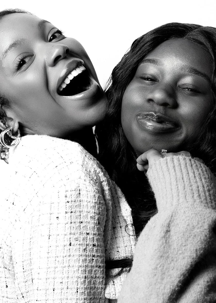 Close-up of two women, one laughing with her mouth open and the other looking at the camera with a slight smile, in black and white.