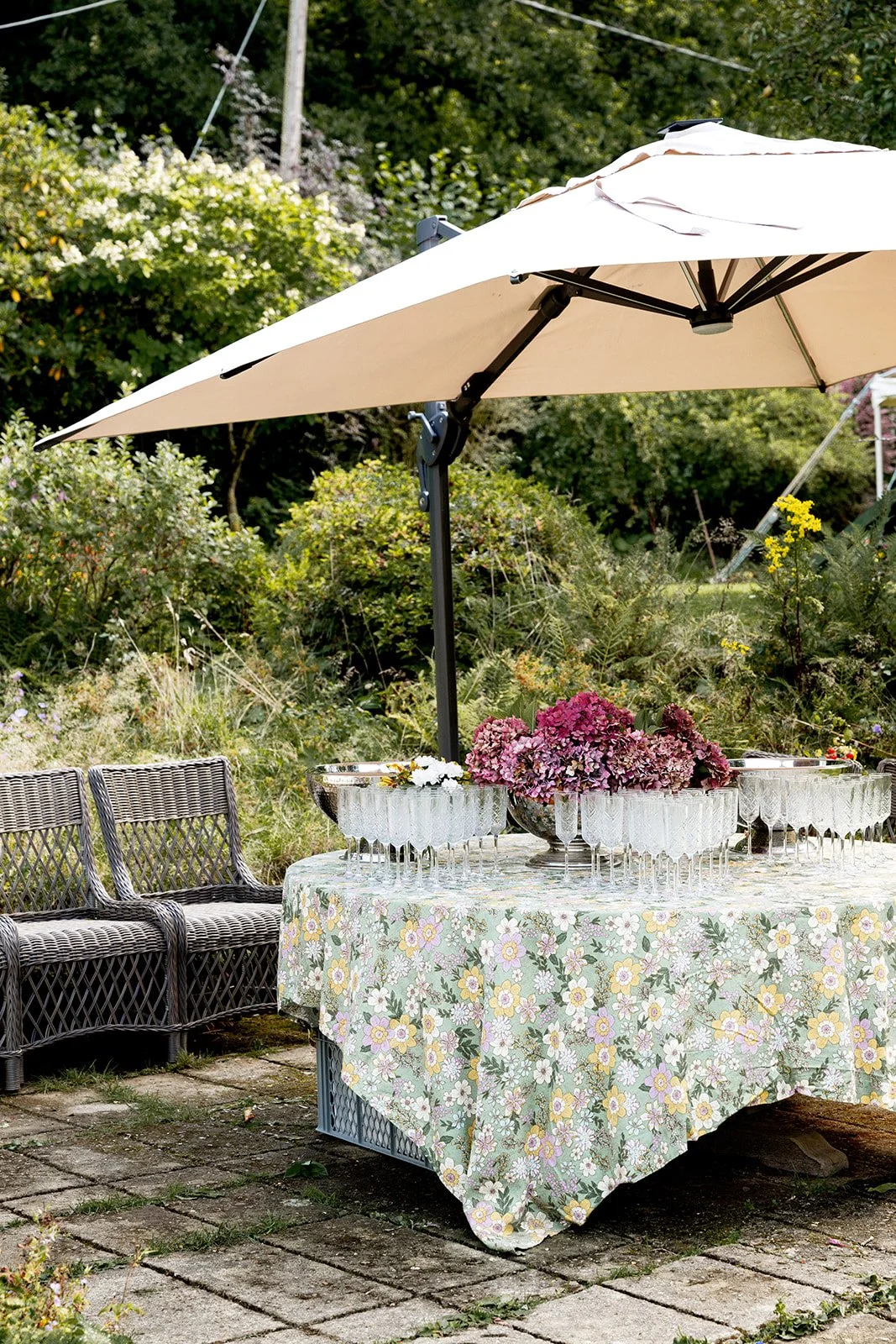Outdoor table setup with a floral tablecloth, glass bowls with pink flowers, and an umbrella in a garden setting