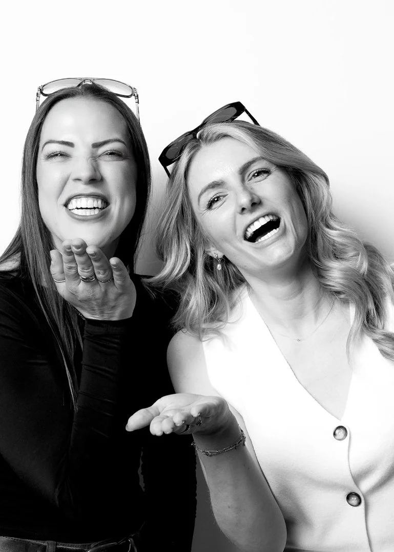 Two women laughing and making playful gestures, wearing sunglasses on their heads, in a black-and-white photo.