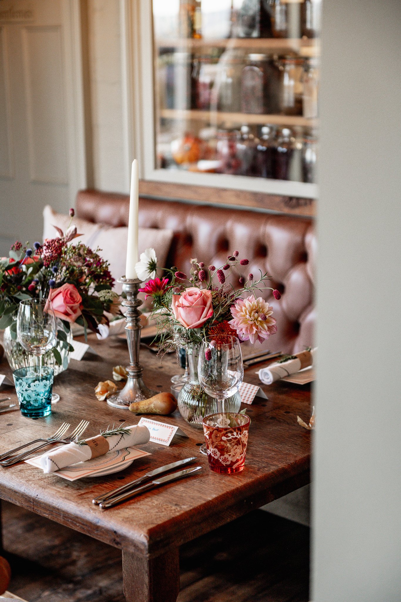 A decorated wooden dining table with floral centerpieces, glassware, cutlery, napkins, and a candle, set for a meal in a cozy room.