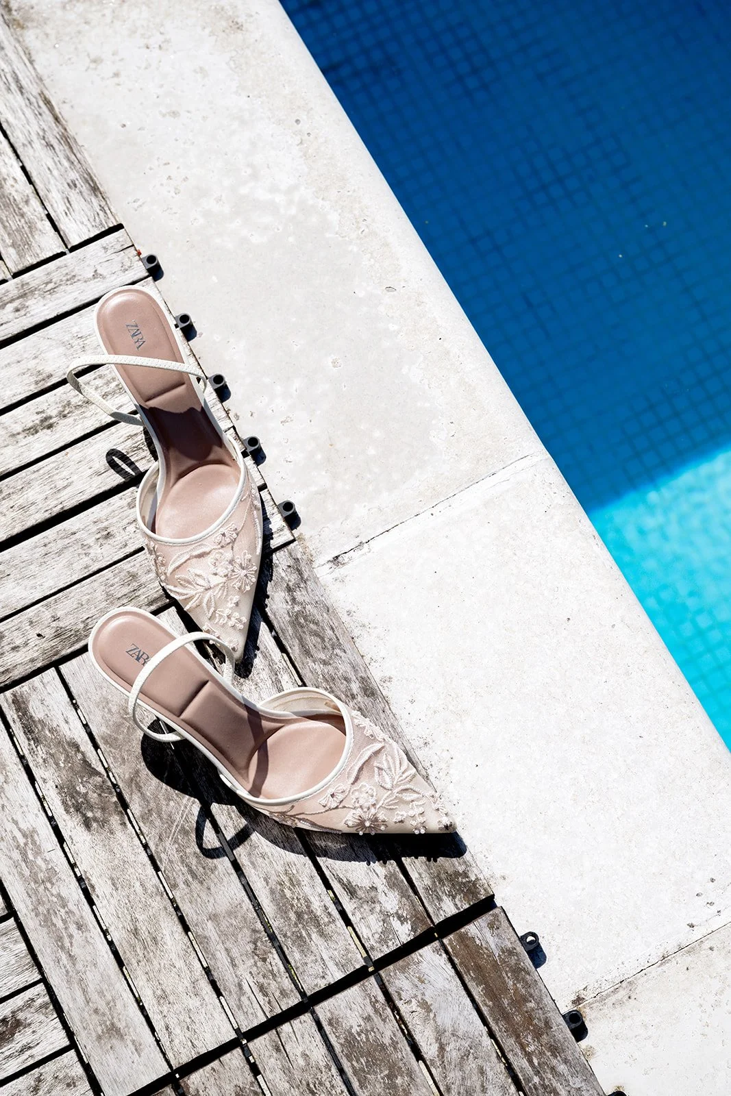 A pair of nude, embroidered high-heeled shoes with ankle straps resting on wooden poolside deck near the edge of a swimming pool.