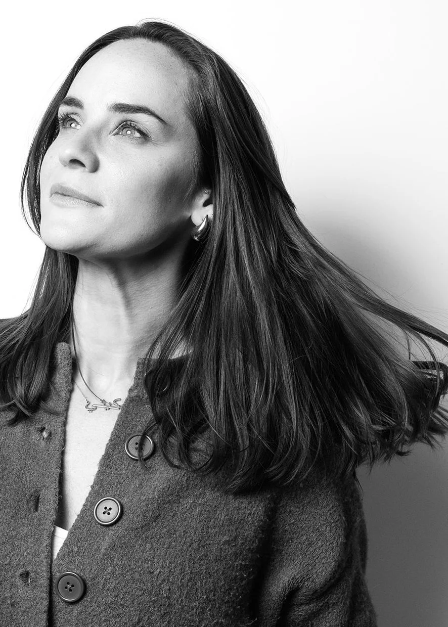 Black and white portrait of a woman with long, dark hair, wearing a coat with large buttons, hoop earrings, and a necklace, looking slightly upward.