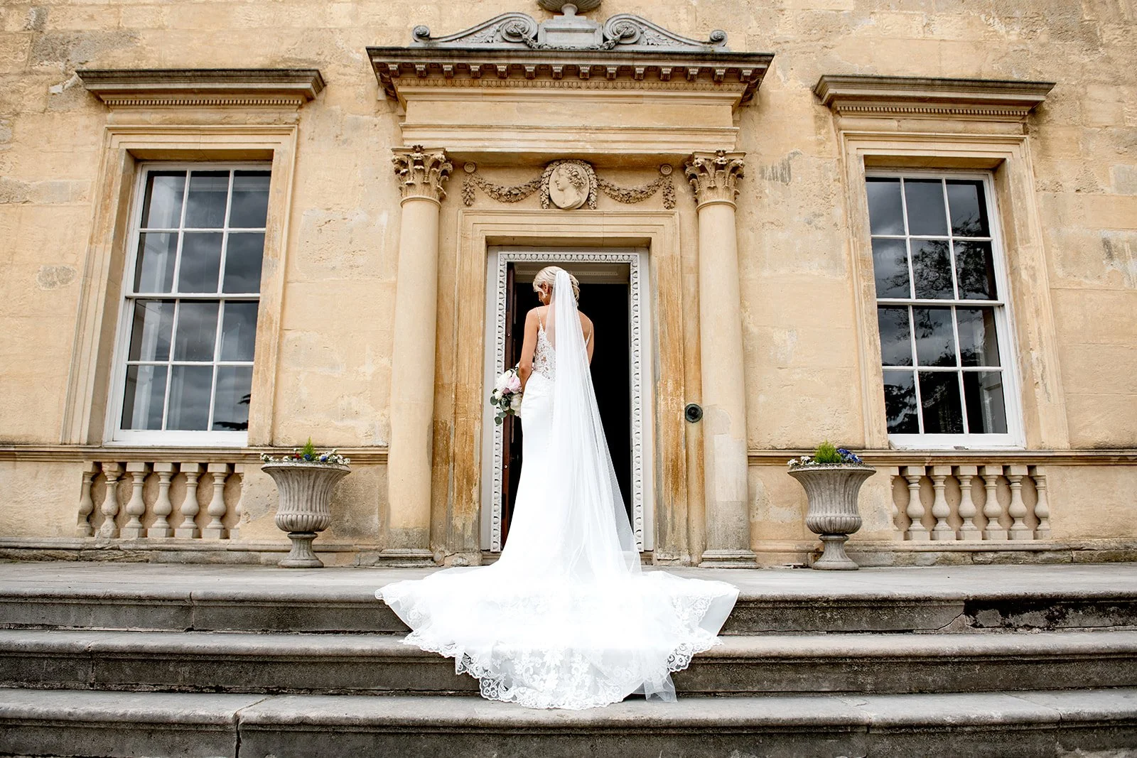 Bride in a white wedding dress with a train and veil standing on stone steps outside a historic building with large windows and decorative architectural details, holding a bouquet of flowers.