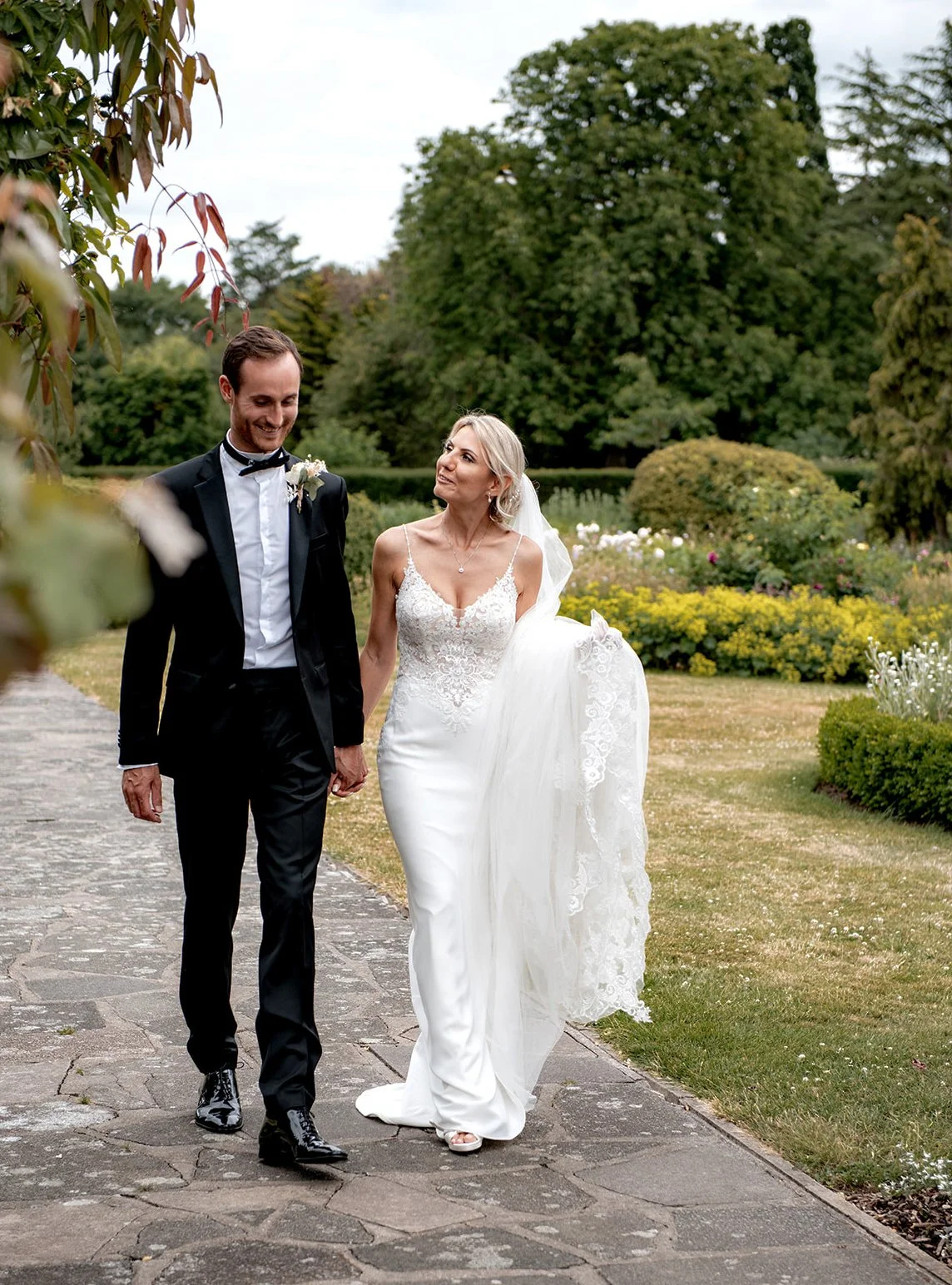 A bride and groom walking hand in hand outdoors on a stone pathway, surrounded by greenery and flowering bushes.