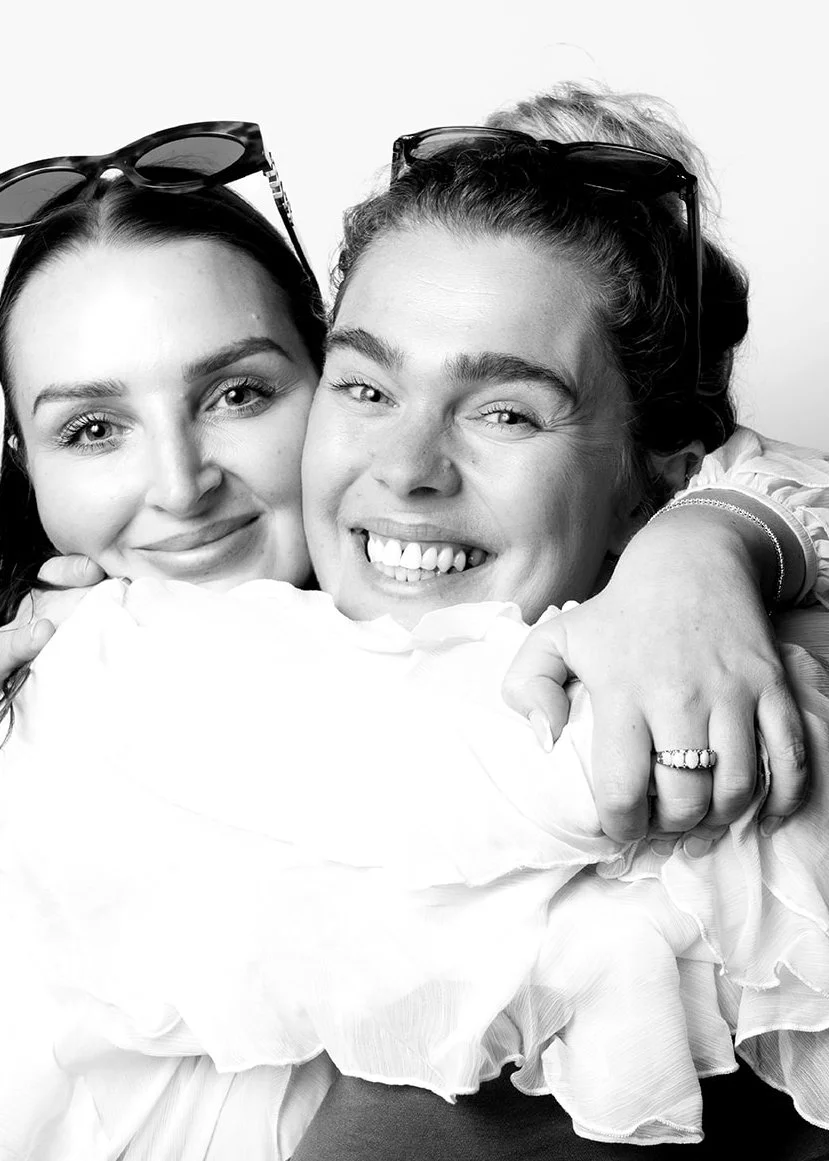 Two women smiling and hugging each other, wearing sunglasses on their heads, in a black and white photo.