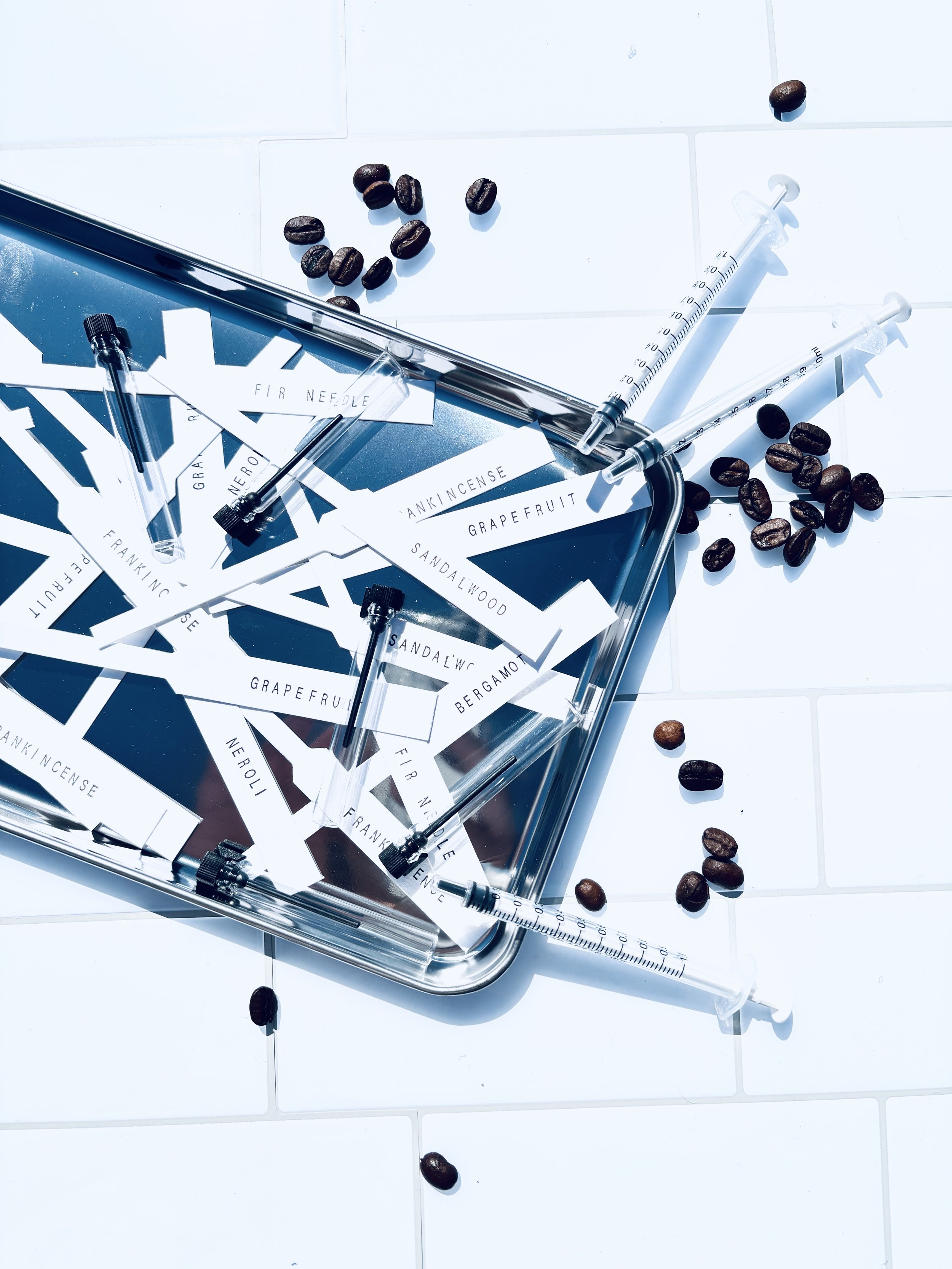 Coffee beans, syringes, and labels for scent notes scattered on a white tiled surface.