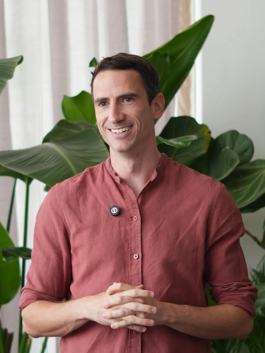 A smiling man with short dark hair wearing a rusty red button-up shirt with sleeves rolled up, standing in front of large green leafy plants near a window with white curtains.
