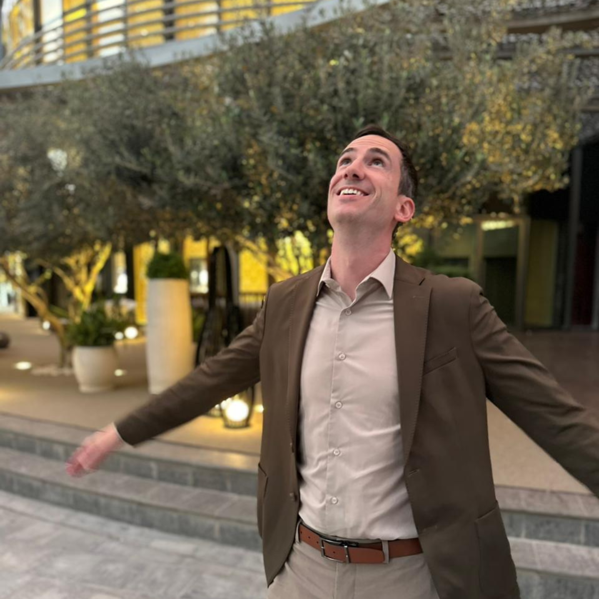 A man in a brown blazer and light shirt outdoors at night with his arms outstretched, smiling and looking upward, with trees and decorative lighting in the background.