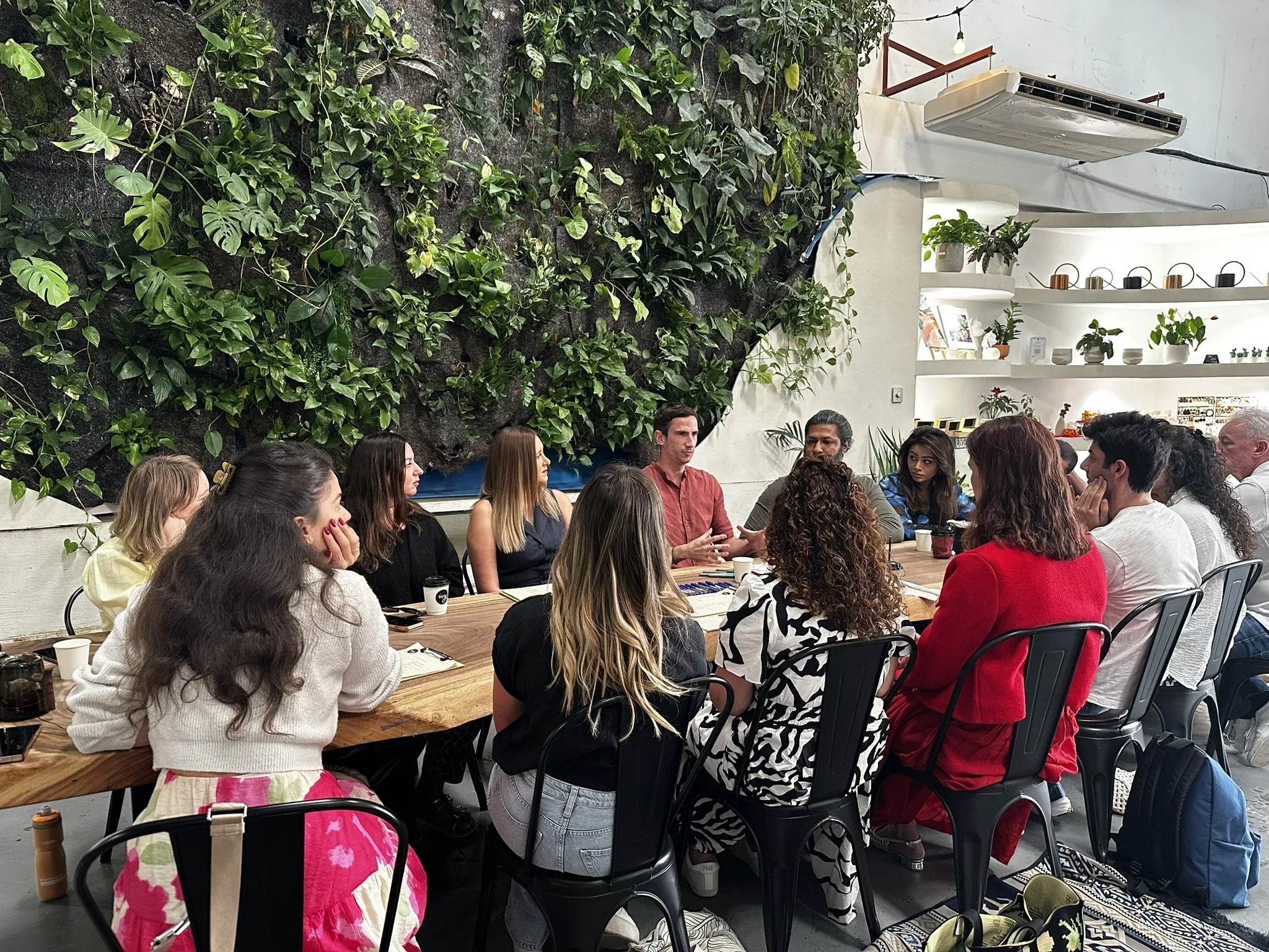 A group of diverse people sitting around a large wooden table in a modern, indoor space with white walls and a large green plant wall. They are engaged in conversation, with some holding cups and notebooks. The space has decorative shelves with plants and cups, and an air conditioning unit on the wall.