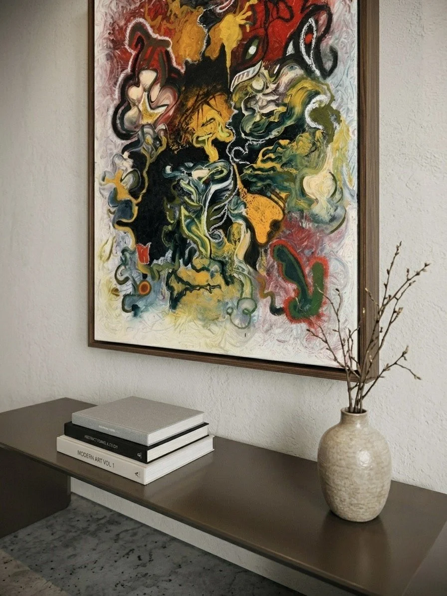 A colorful abstract painting hangs on a cream-colored wall above a dark wooden sideboard, which holds stacked books and a beige vase with bare branches.