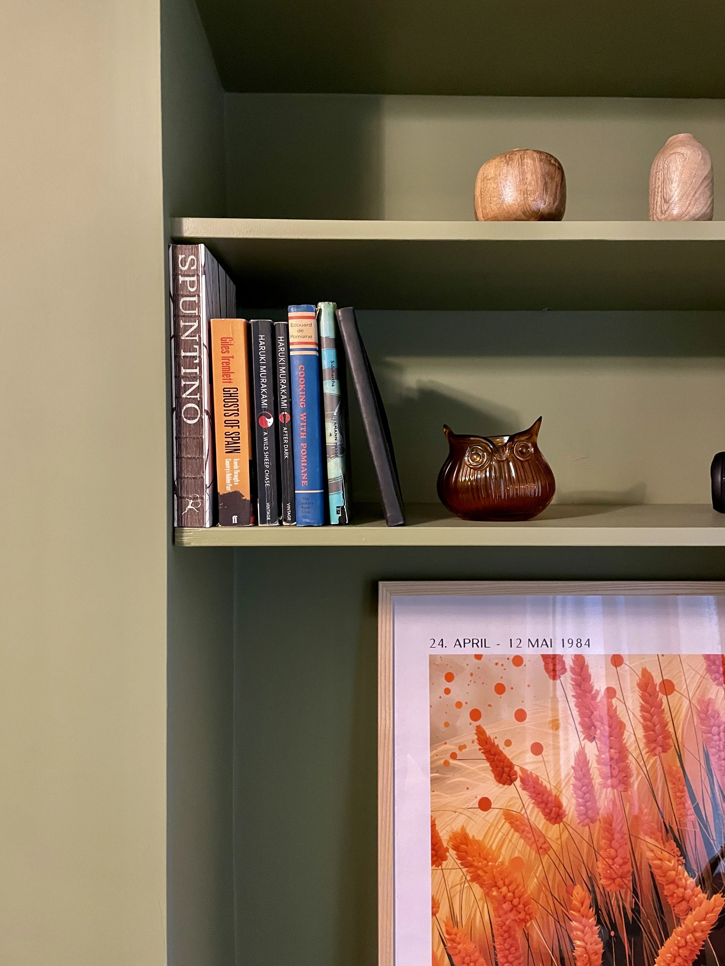 Green bookshelf with decorative wooden objects at the top, a row of books including 'Spuintino,' 'Ghosts of Spain,' and 'Cooking with Pomegranate,' a brown owl-shaped vase, and a framed artwork featuring a floral design with orange and pink hues and 