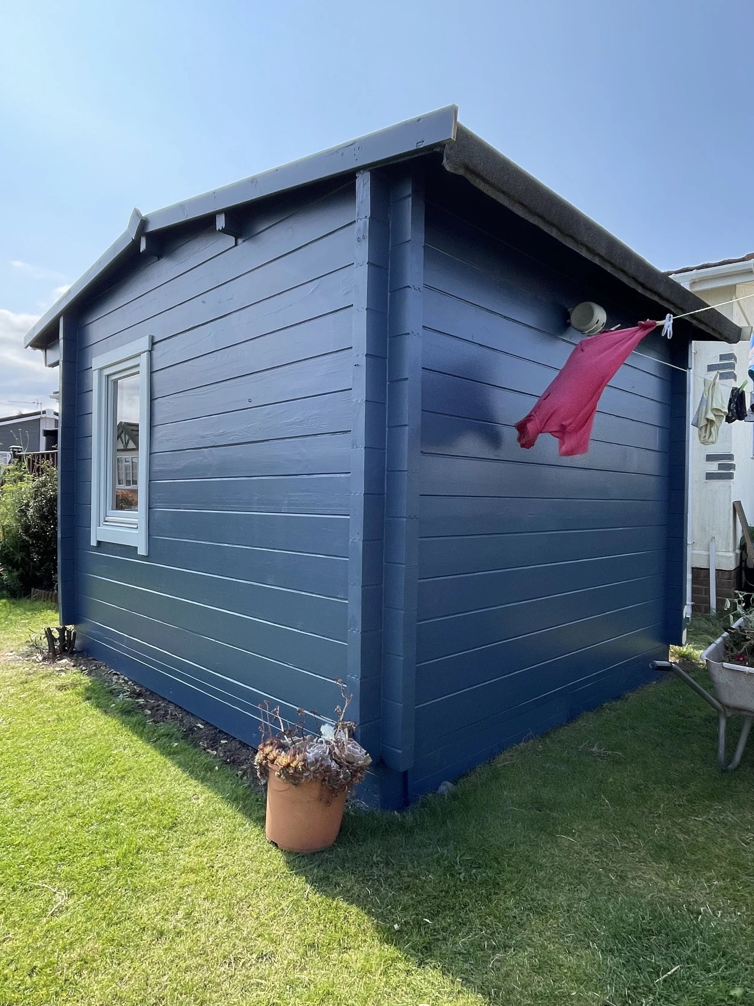 A small blue wooden shed with a window, located in a backyard with green grass, potted plants, a wheelbarrow, and laundry hanging on a line.