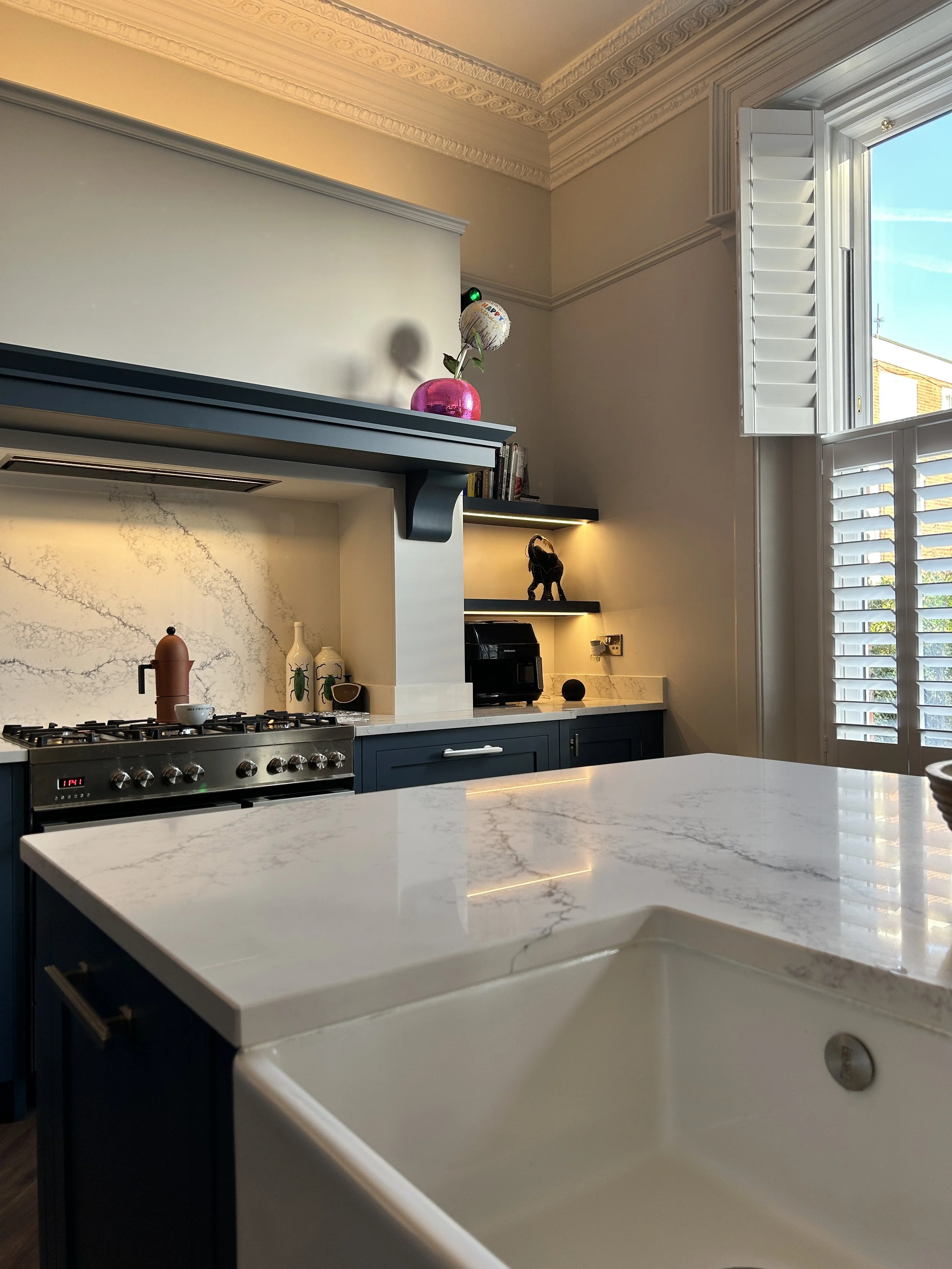 Modern kitchen with blue cabinets, white marble countertops, and a kitchen island. There is a stove with a coffee pot on it, decorative vases, a black appliance, and open shelves with a small sculpture of a dog. A large window with white shutters all