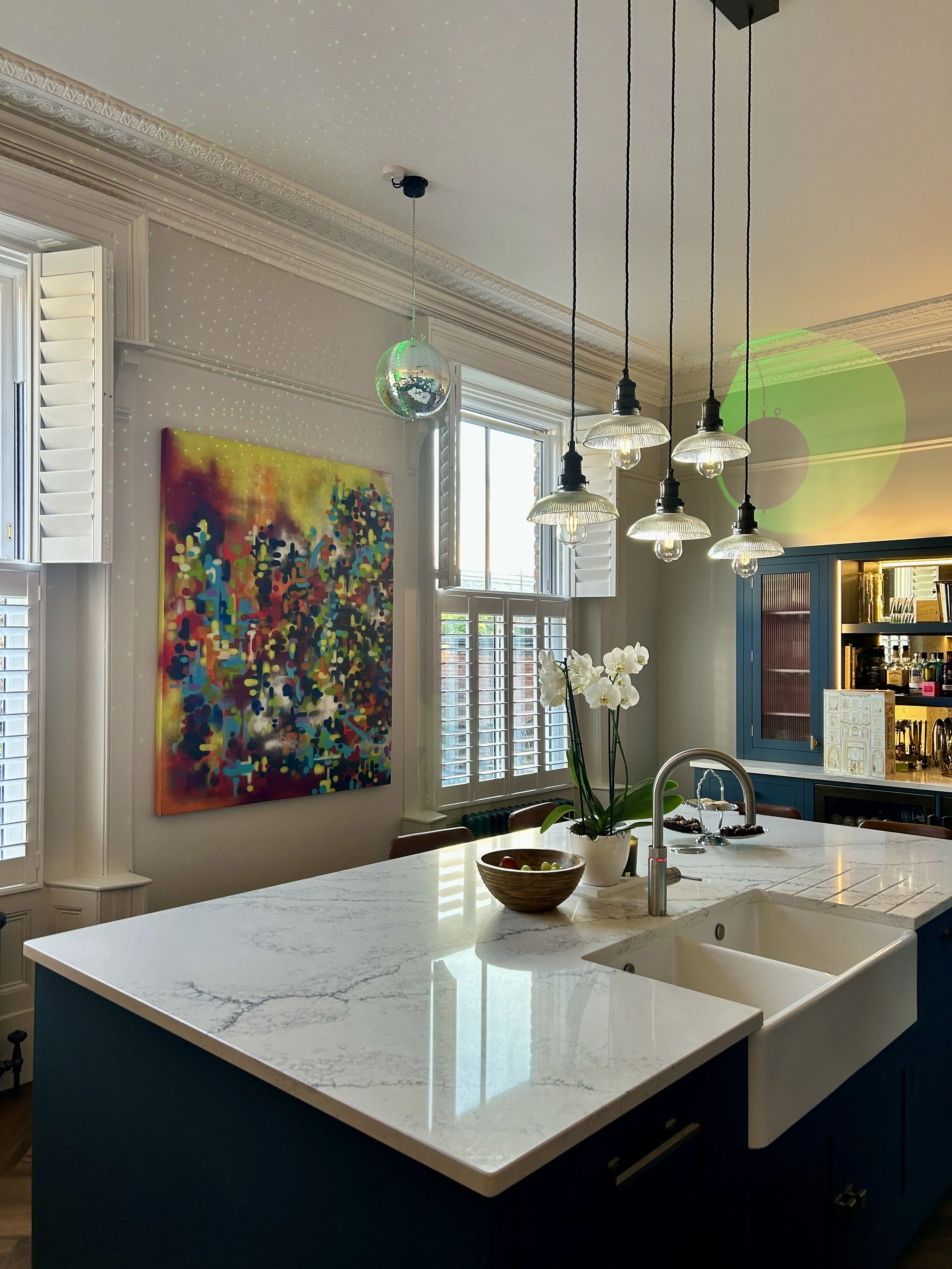 Modern kitchen with white marble island, white sink, blue cabinetry, colorful abstract painting on the wall, pendant lights hanging from ceiling, and orchid plant on the countertop.