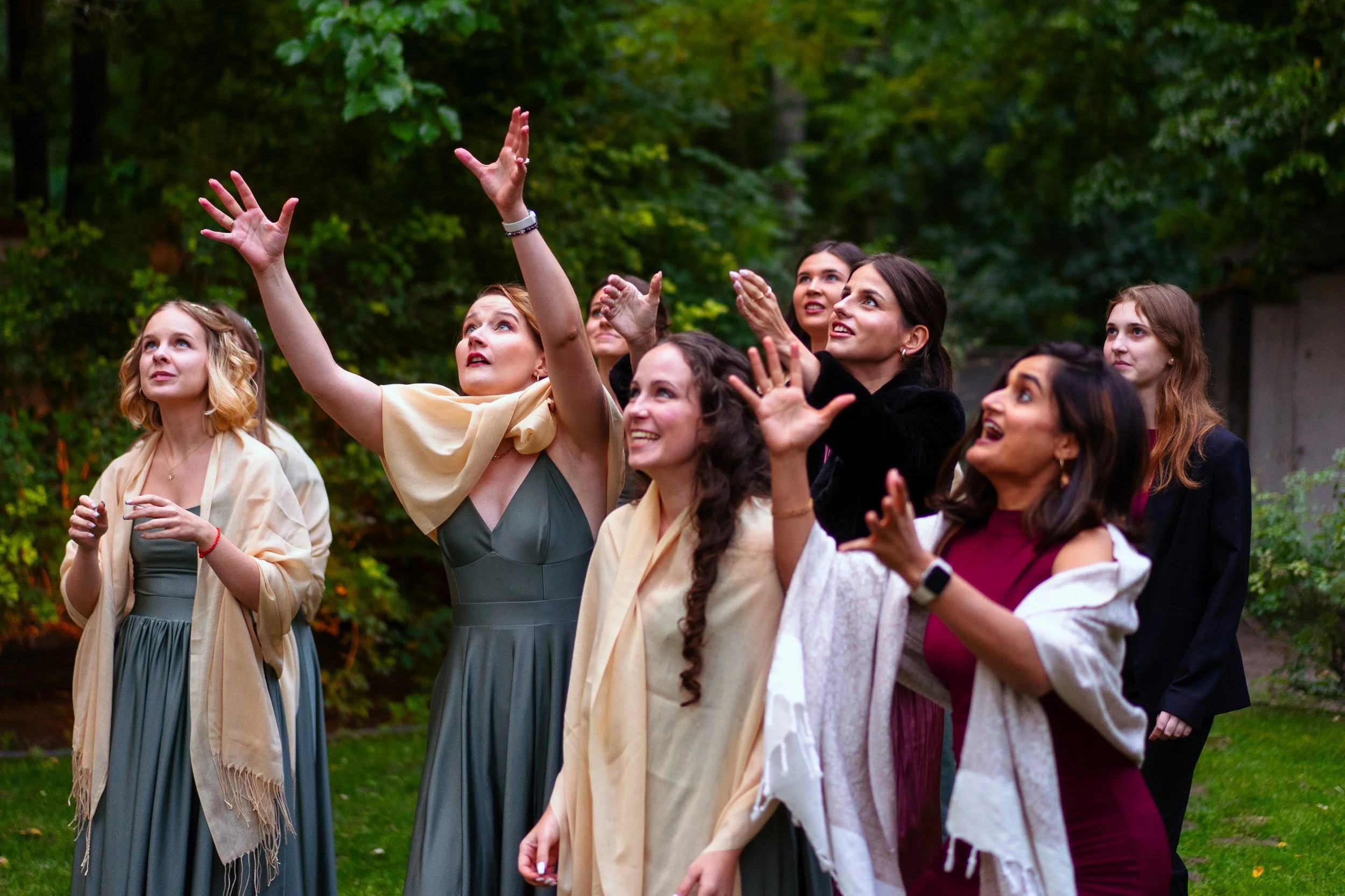 A group of women outdoors, celebrating and reaching towards the sky with joyful expressions, surrounded by greenery.