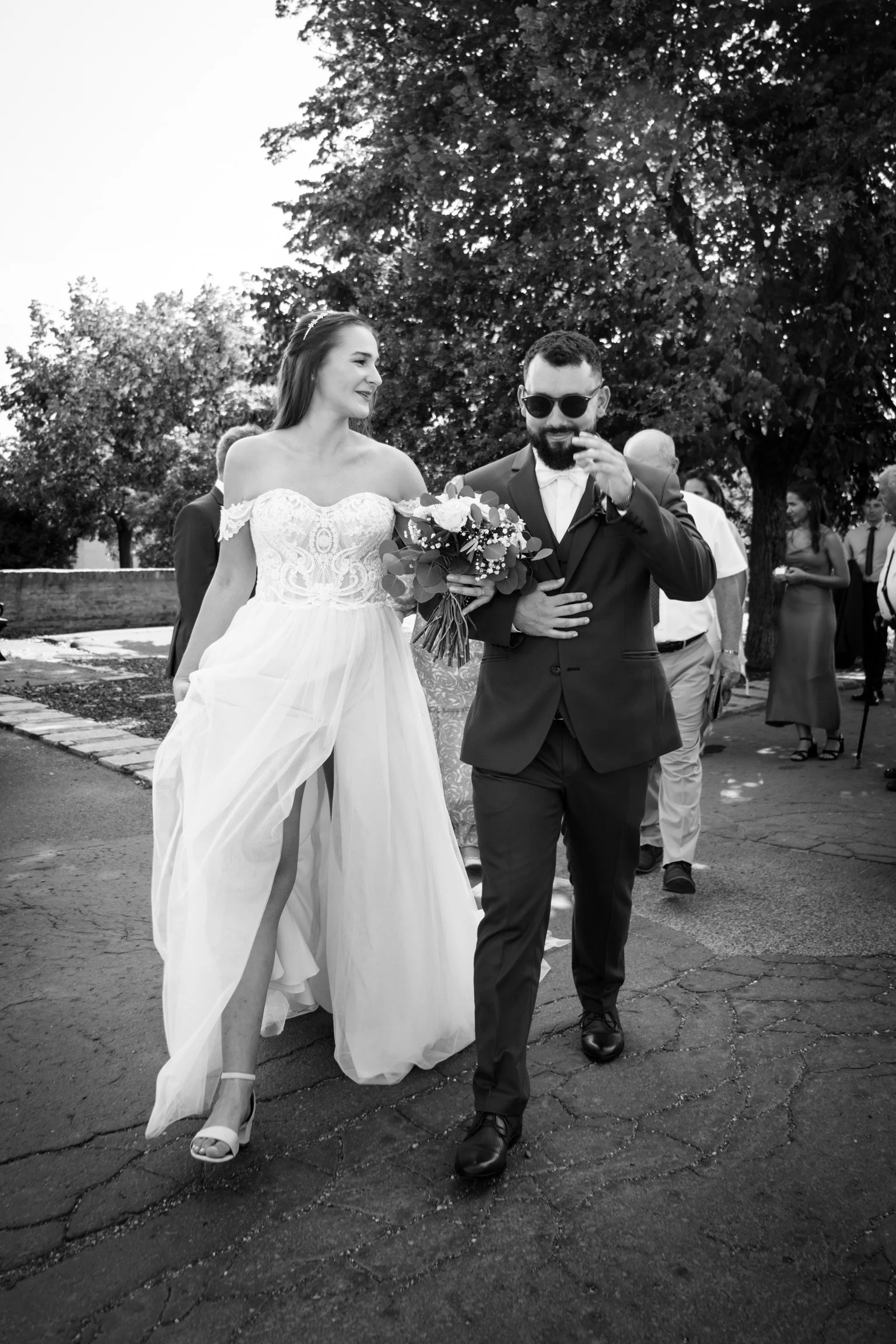A bride in a wedding dress holding a bouquet walking beside a groom in a suit with sunglasses, surrounded by wedding guests outdoors.