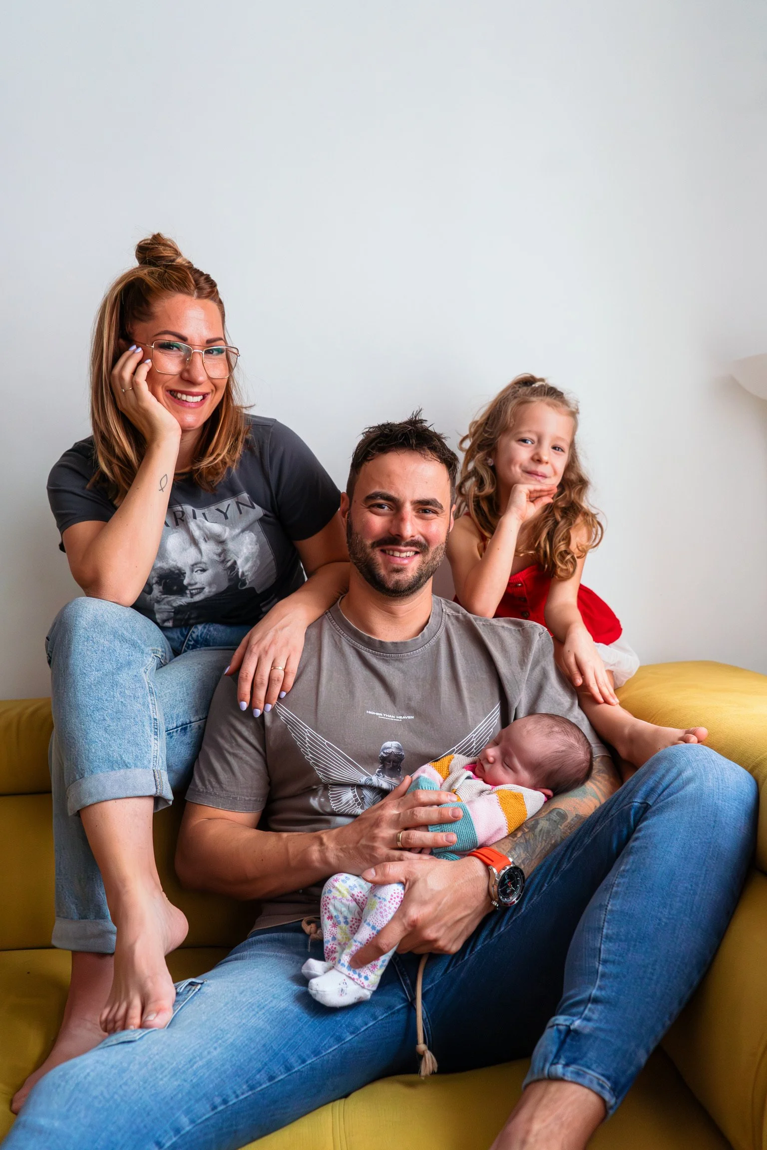 Lifestyle otthoni családi fotózás Hódmezővásárhelyen: négyszemélyes család mosolyog egy sárga kanapén, az édesapa karjaiban egy békésen alvó újszülött kisbabával.