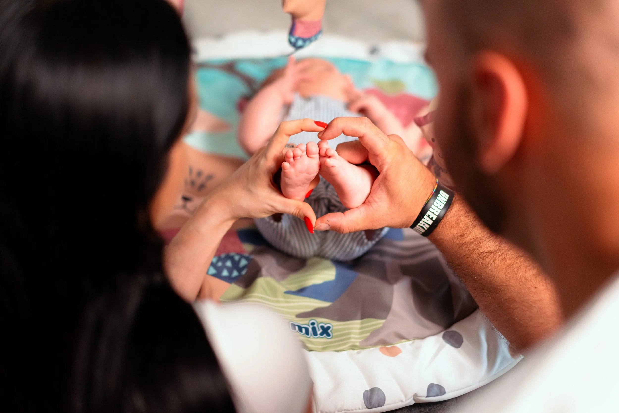 Close-up of a newborn baby held in a baby's bath with support from multiple adults, focused on tiny feet forming a heart shape with their hands.