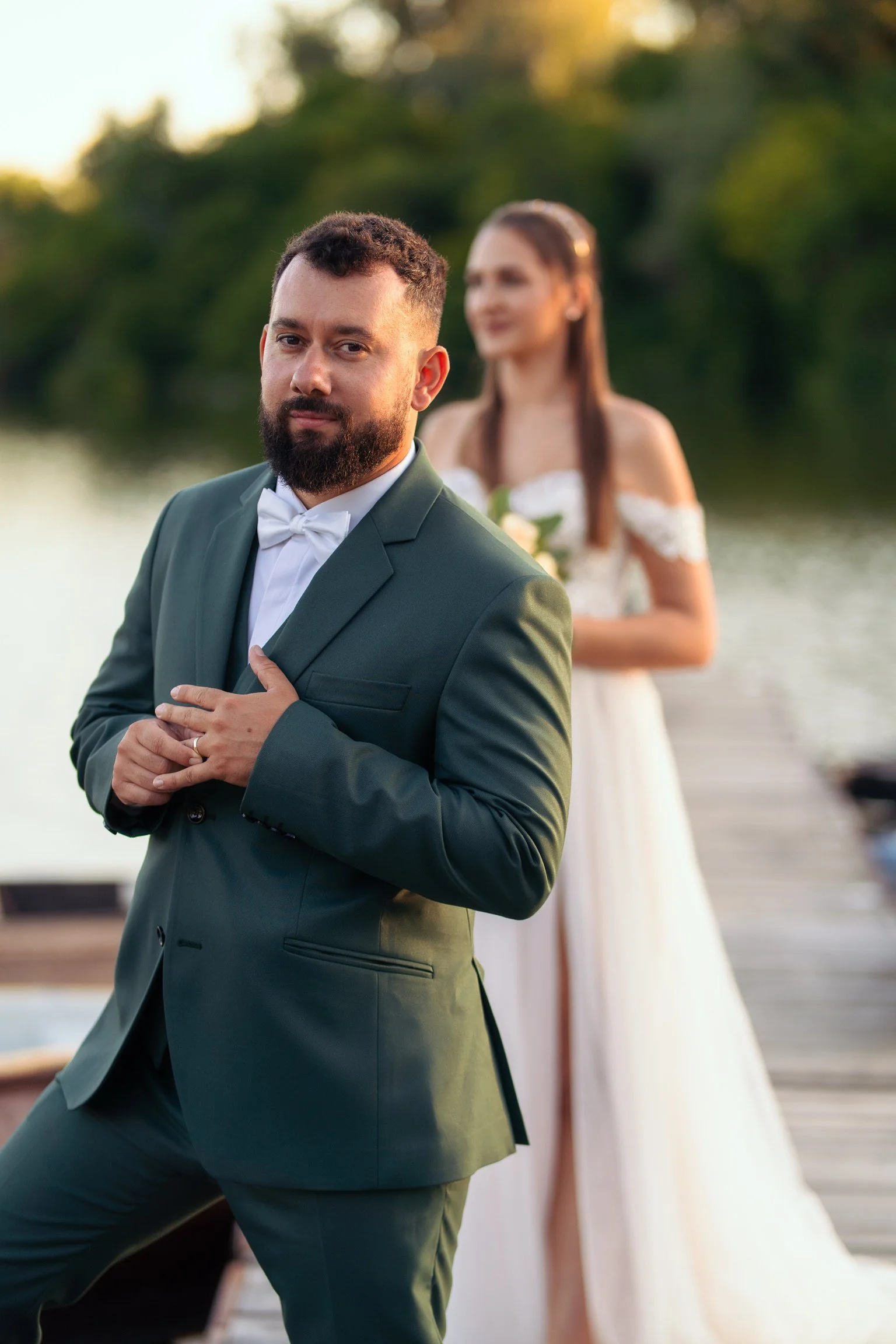 Kreatív esküvői fotó a Tisza-parti stégen Szeged környékén: előtérben a stílusos zöld öltönyös vőlegény igazítja gyűrűjét, a háttérben életlenül a menyasszony látható.