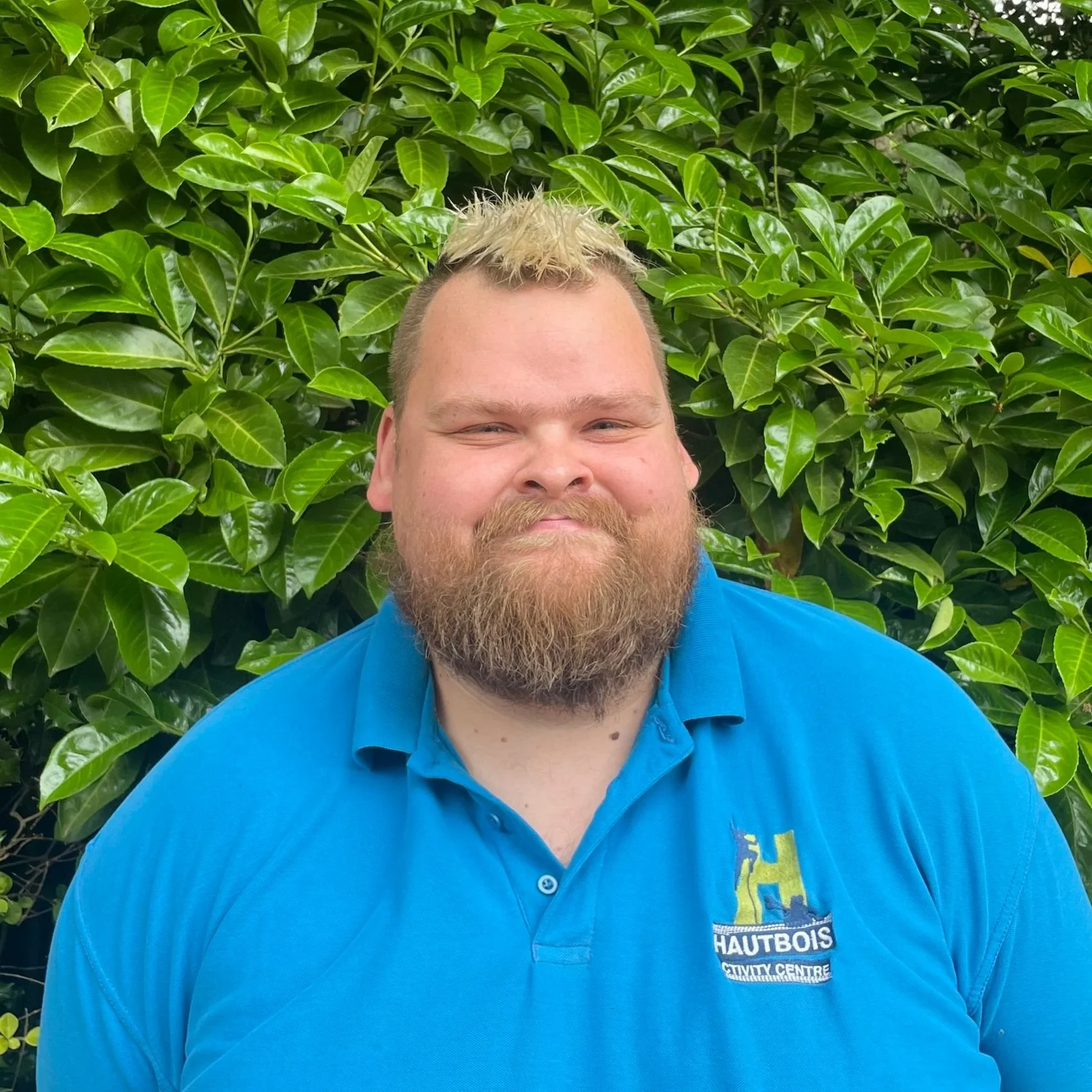 Portrait of a man with short brown hair, smiling, wearing a bright blue shirt, standing in front of green foliage.
