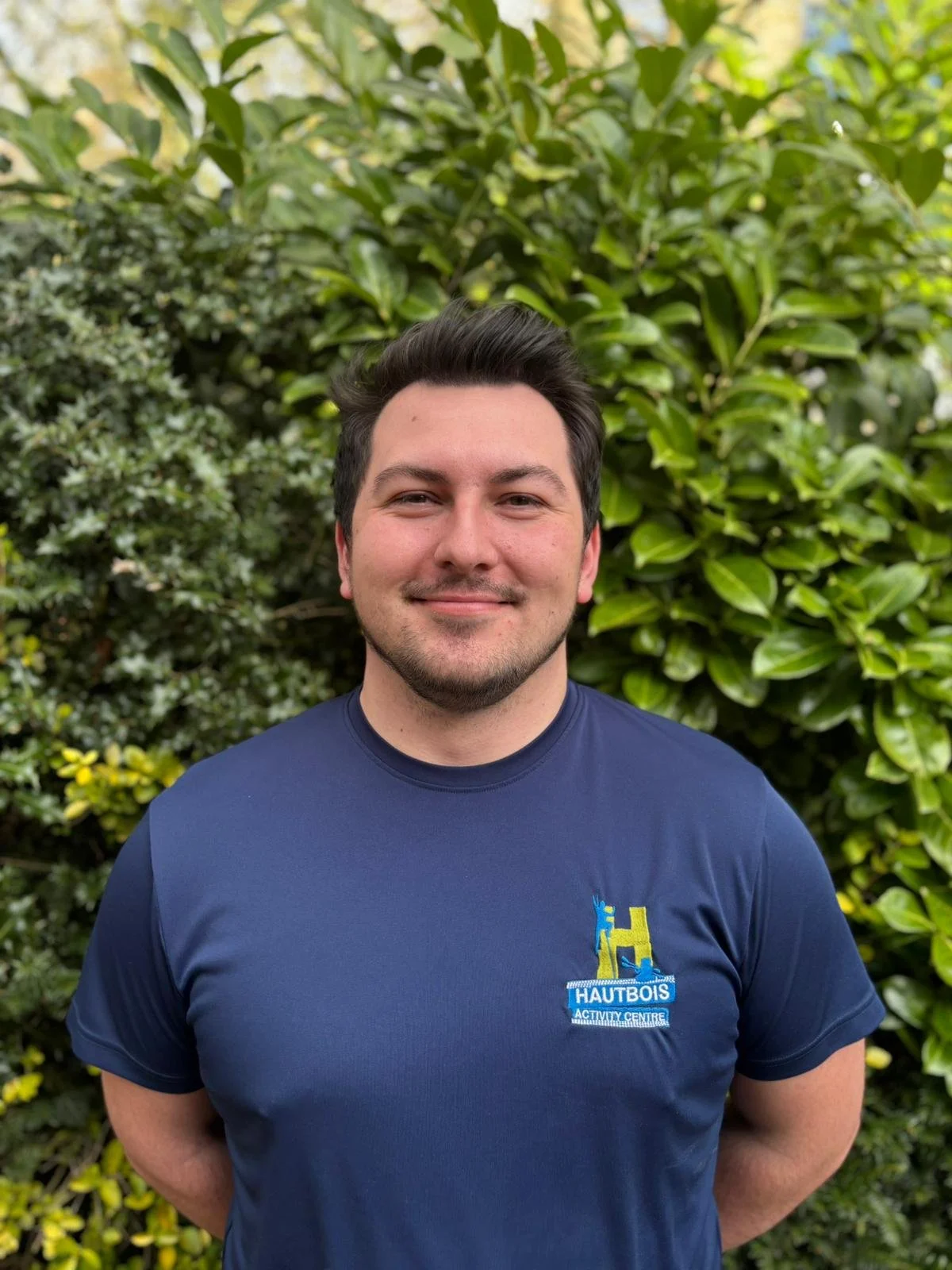A young man smiling outdoors, standing in front of green foliage, wearing a dark fleece jacket and a blue collared shirt.