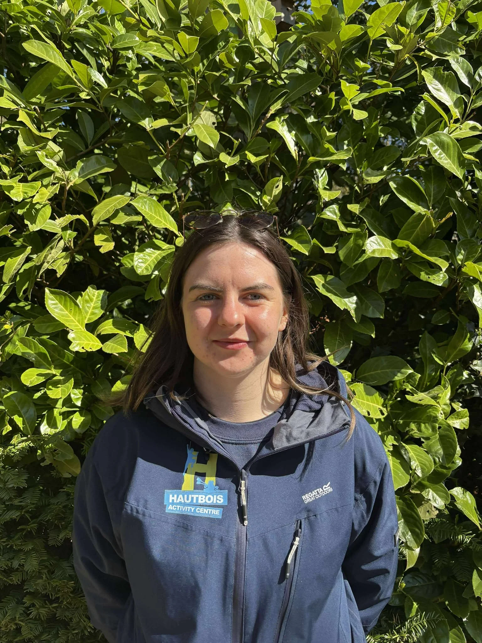 A young woman with long red hair and glasses smiling outdoors in front of green foliage, wearing a blue shirt.