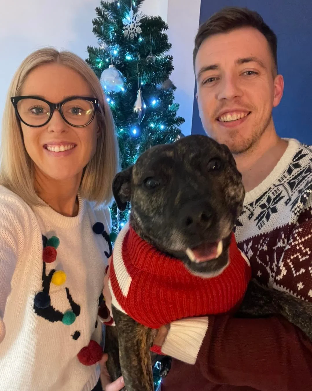 A couple with a dog in front of a Christmas tree, all wearing holiday sweaters, smiling for a photo.