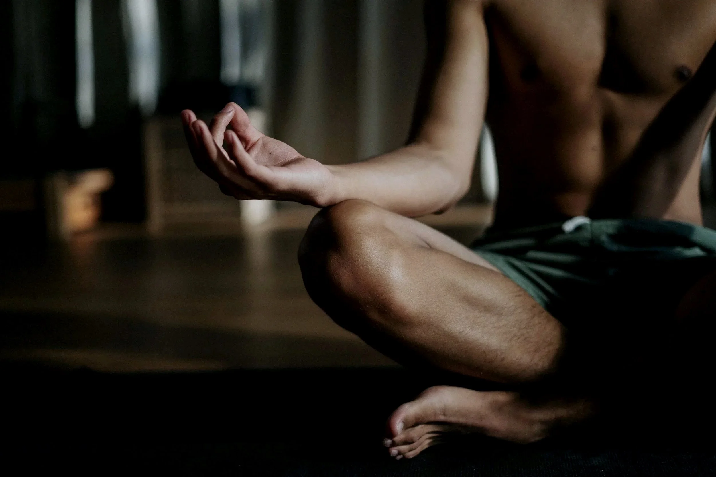 A person practicing yoga or meditation indoors, sitting cross-legged with one hand resting on their knee and fingers in a mudra gesture, wearing only shorts, with a dark and blurred background.