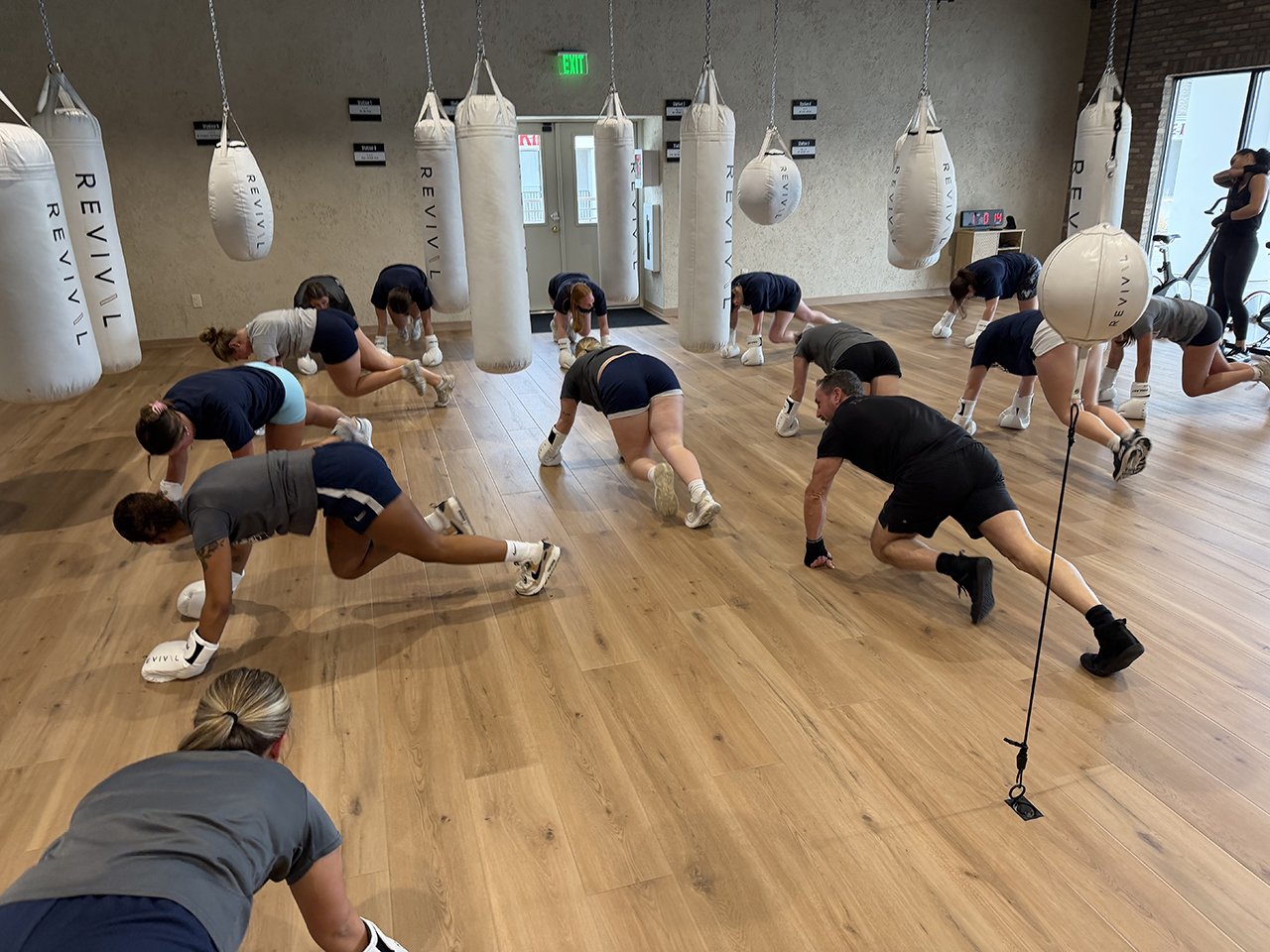 People participating in a group fitness class at a gym with punching bags. They are performing a mountain climbers exercise on the wooden floor.