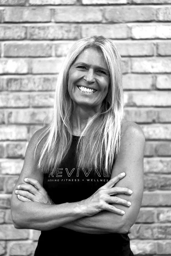 A smiling woman with long blonde hair, wearing a sleeveless top, standing with arms crossed in front of a brick wall.