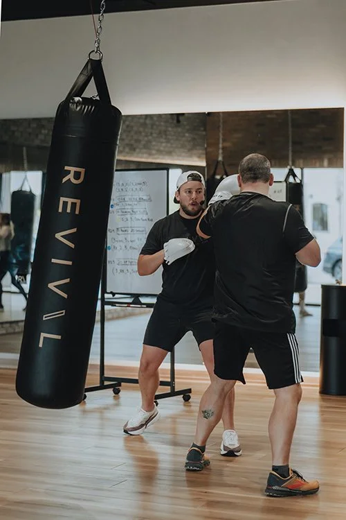 Two men training in the REVIVAL fitness studio, one wearing gloves and the instructor wearing mits facing him, with a punching bag labeled 'REVIVAL' hanging nearby.