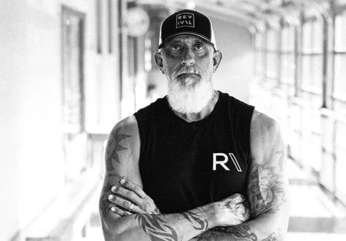 Black and white photo of a man with a beard and tattoos, wearing a sleeveless shirt and a cap, standing with arms crossed in a corridor.