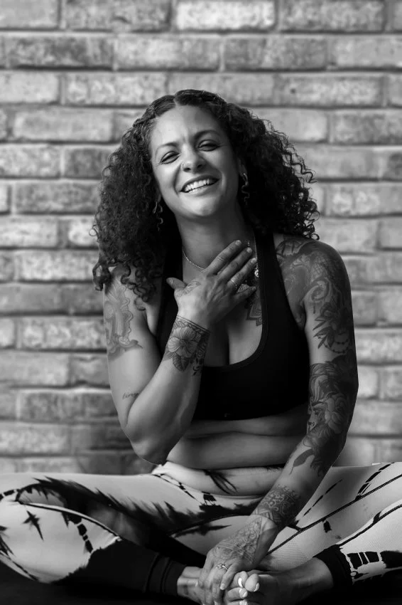 A smiling woman with curly hair, tattoos on her arms, and wearing a sports bra and patterned leggings, sitting cross-legged against a brick wall.