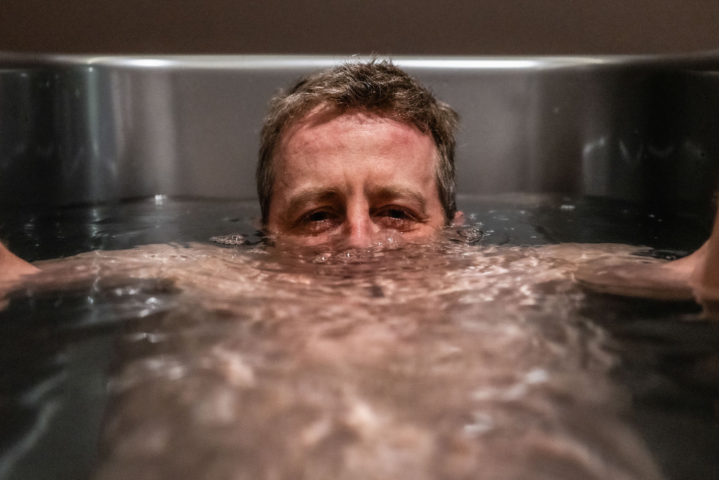 A person with short hair is submerged in water up to their eyebrows, in a metallic or stainless steel container or sink.