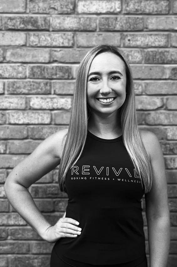 A woman with long, straight hair smiling at the camera, wearing a sleeveless top that says "Reviv" and has the words "Boxing Fitness Wellness" underneath, standing against a brick wall.