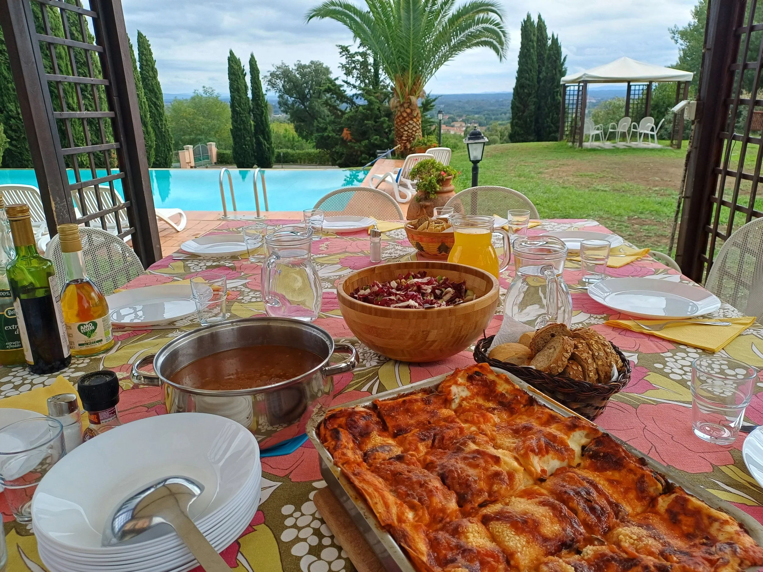 Outdoor dining table set with pizza, salad, bread, and drinks, overlooking a swimming pool and lush greenery on a cloudy day.