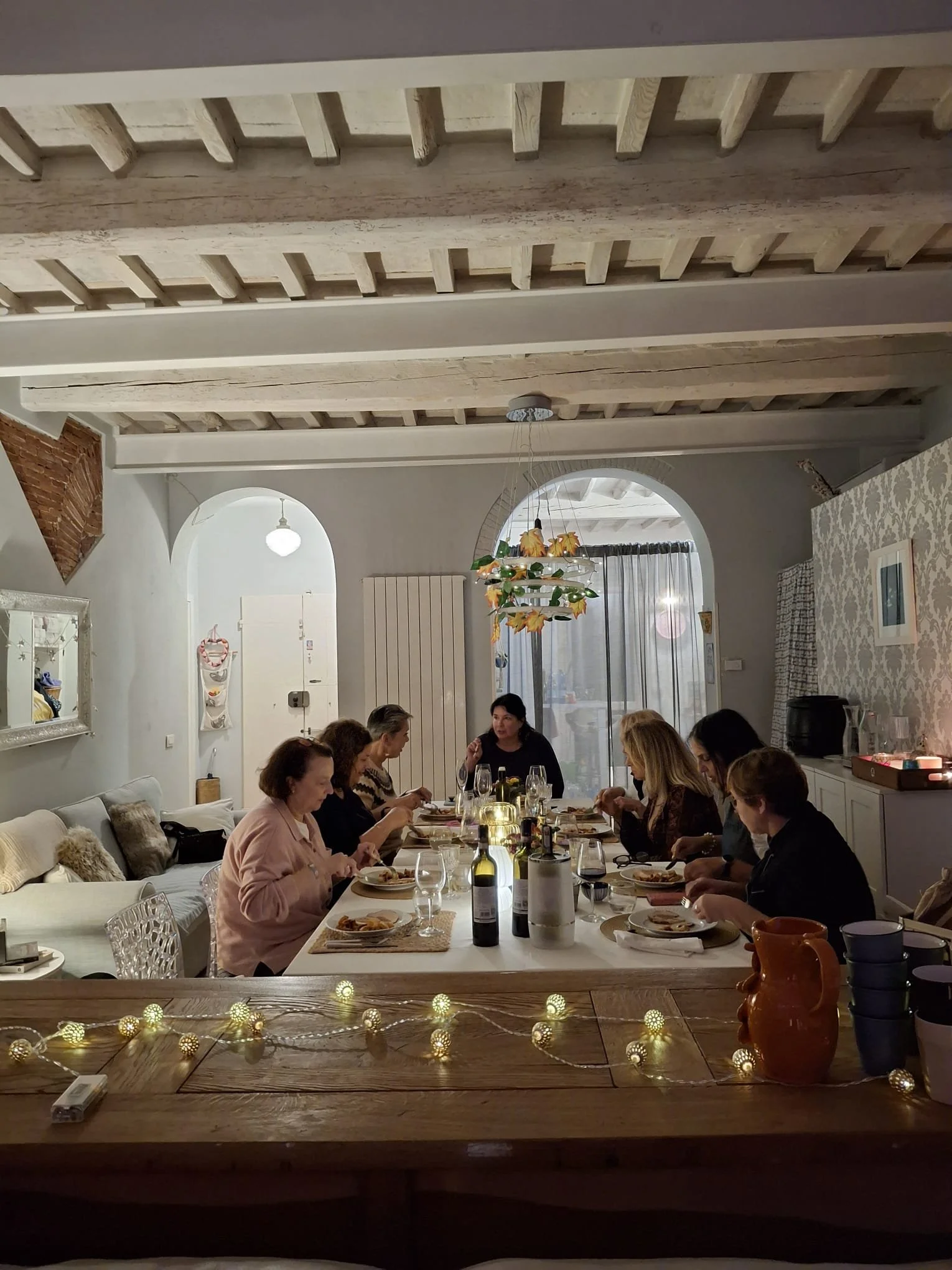 Group of women having dinner around a long table in a cozy, decorated dining room with string lights, bottles of wine, and candles.