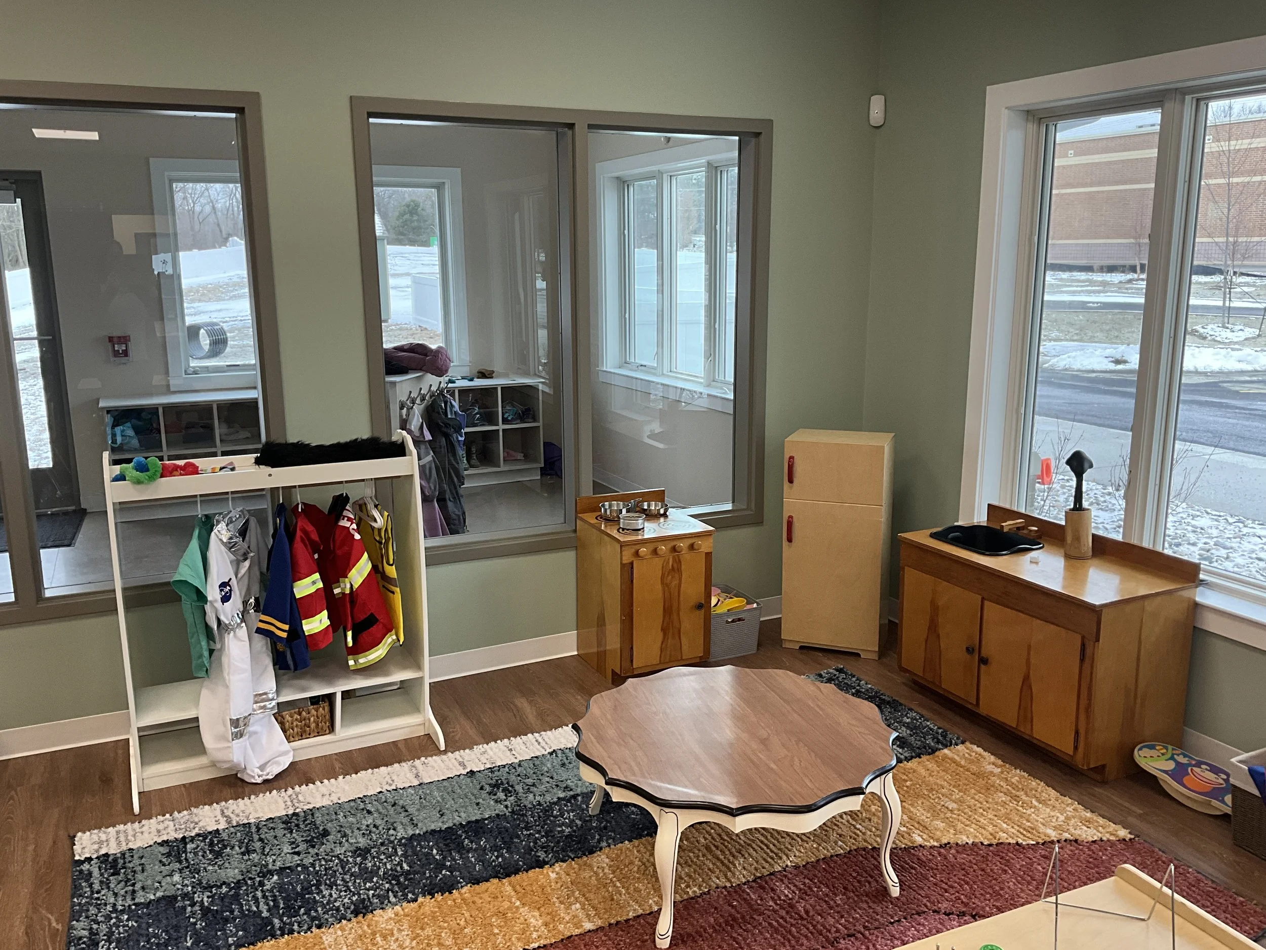 Preschool classroom with wooden furniture, a colorful rug, and a coat rack with play clothes. Windows show snow outside.