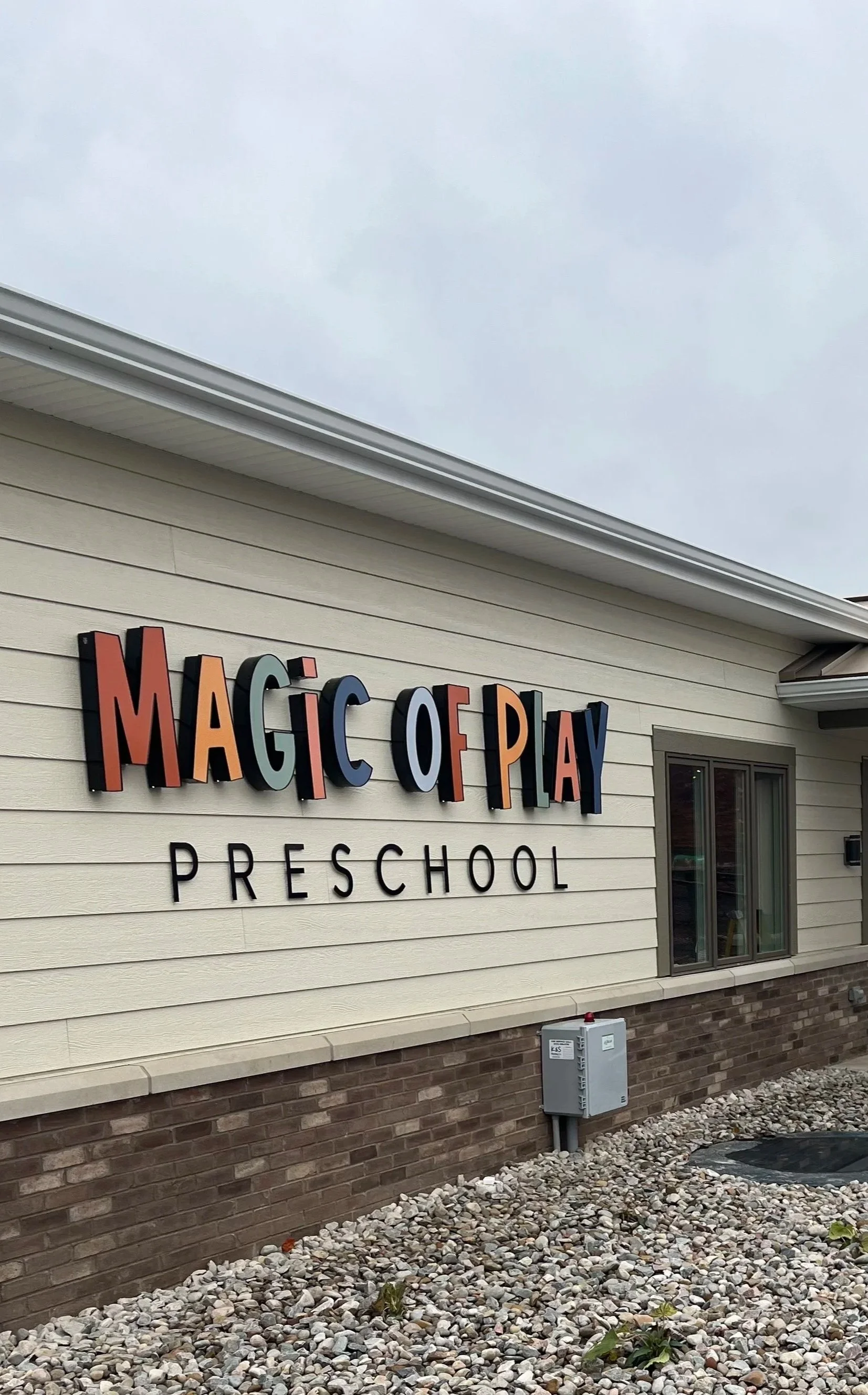 Exterior view of Magic of Play Preschool with colorful 3D sign on siding, window, and gravel landscaping