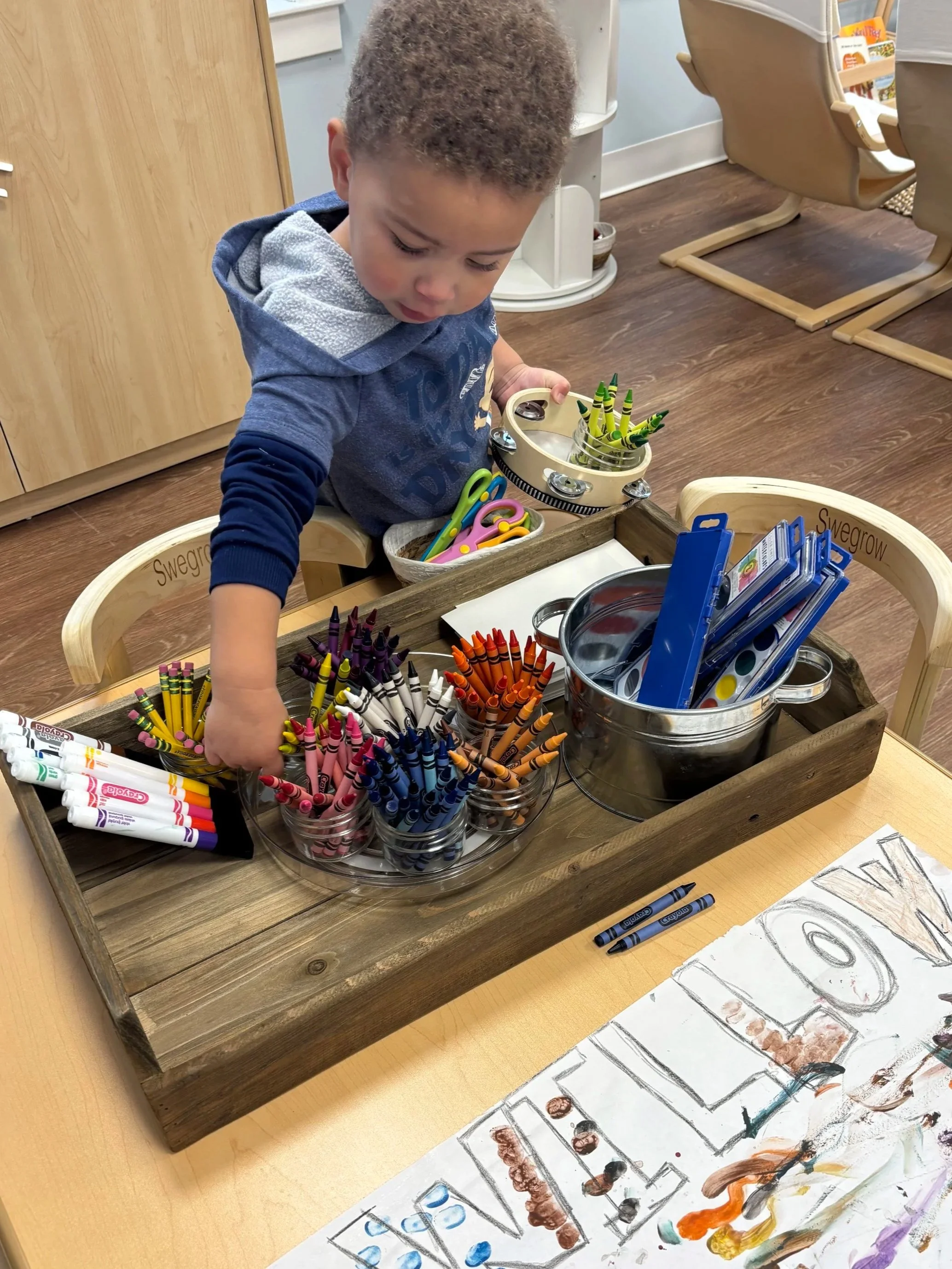 A young child reaching for colored crayons in glass jars on a wooden tray, with drawing supplies and artwork on the table in a classroom setting.