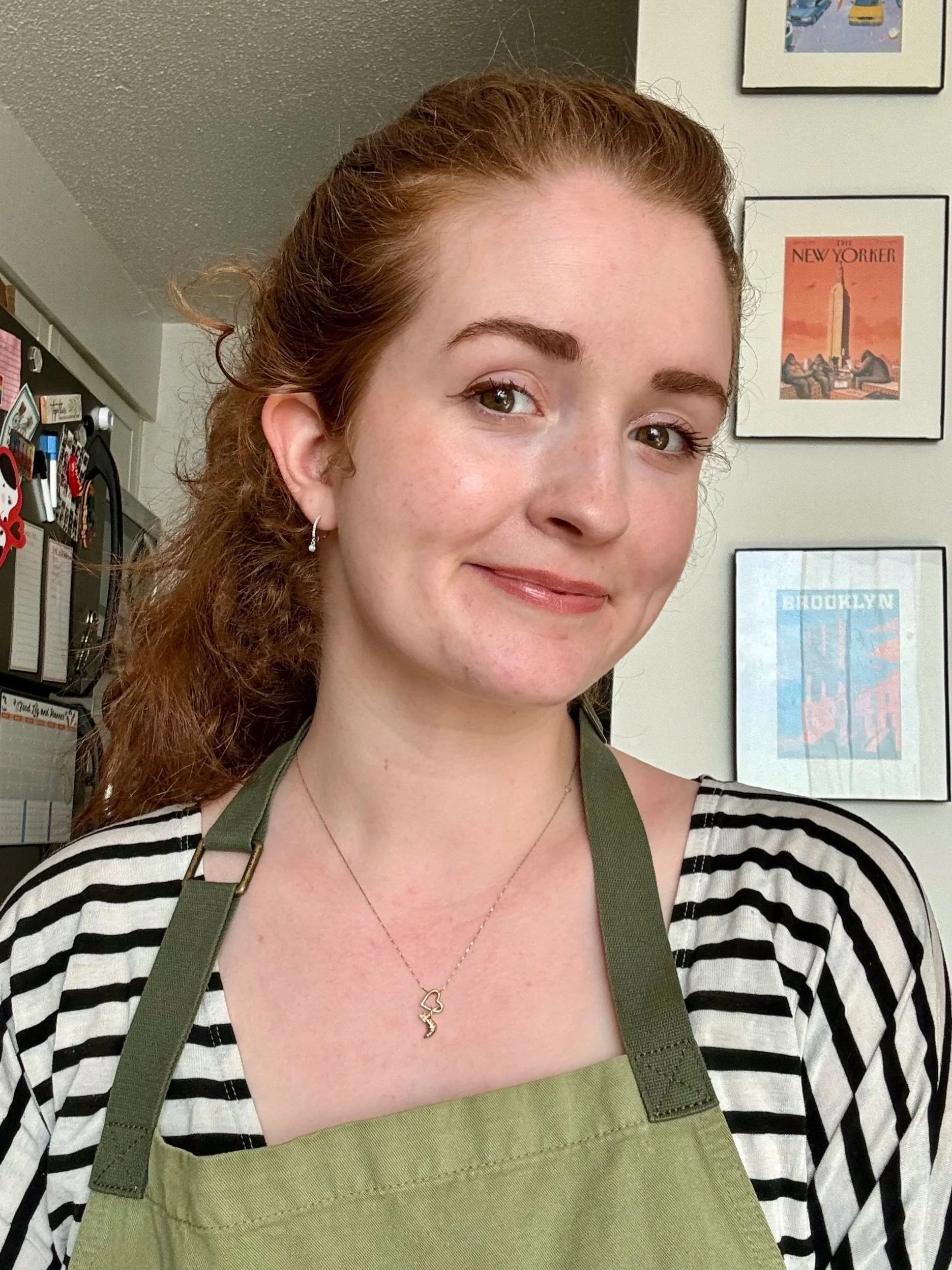 A woman with red hair, wearing a striped shirt, a green apron, and a necklace with a pendant, smiling indoors with framed posters on the wall behind her.