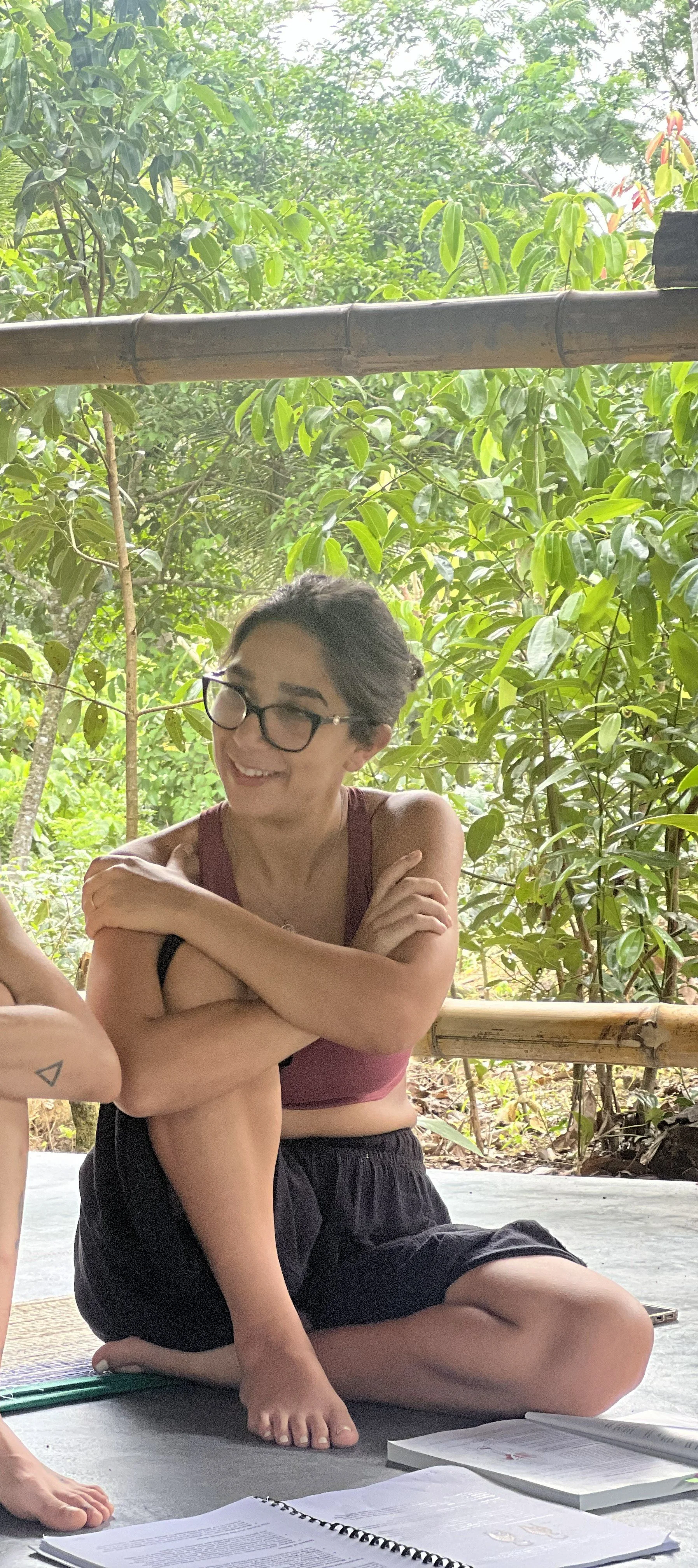 Woman with glasses sitting on the ground in a lush outdoor setting, smiling with arms crossed over knees, surrounded by green foliage.