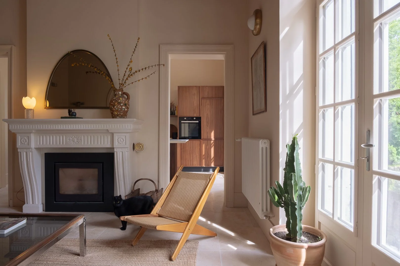 Intérieur d'un salon lumineux avec cheminée blanche décorée d'une grande vase en osier avec des branches, un miroir semi-circulaire, une lampe, une plante cactus, un fauteuil en bois et ratan, et une porte-fenêtre laissant passer la lumière naturelle.