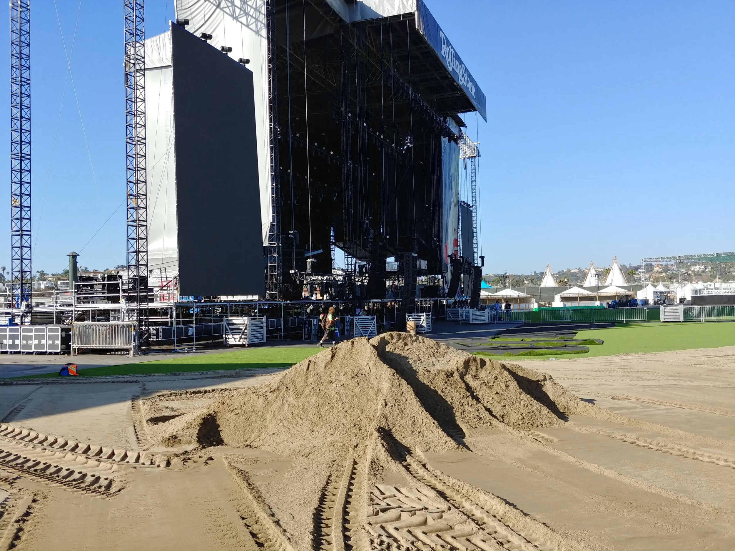 A large outdoor stage setup at a music festival with a giant screen on the left and black scaffolding supporting lighting and sound equipment. In the foreground, there is a large pile of dirt with tire tracks. The sky is clear and there are tents and