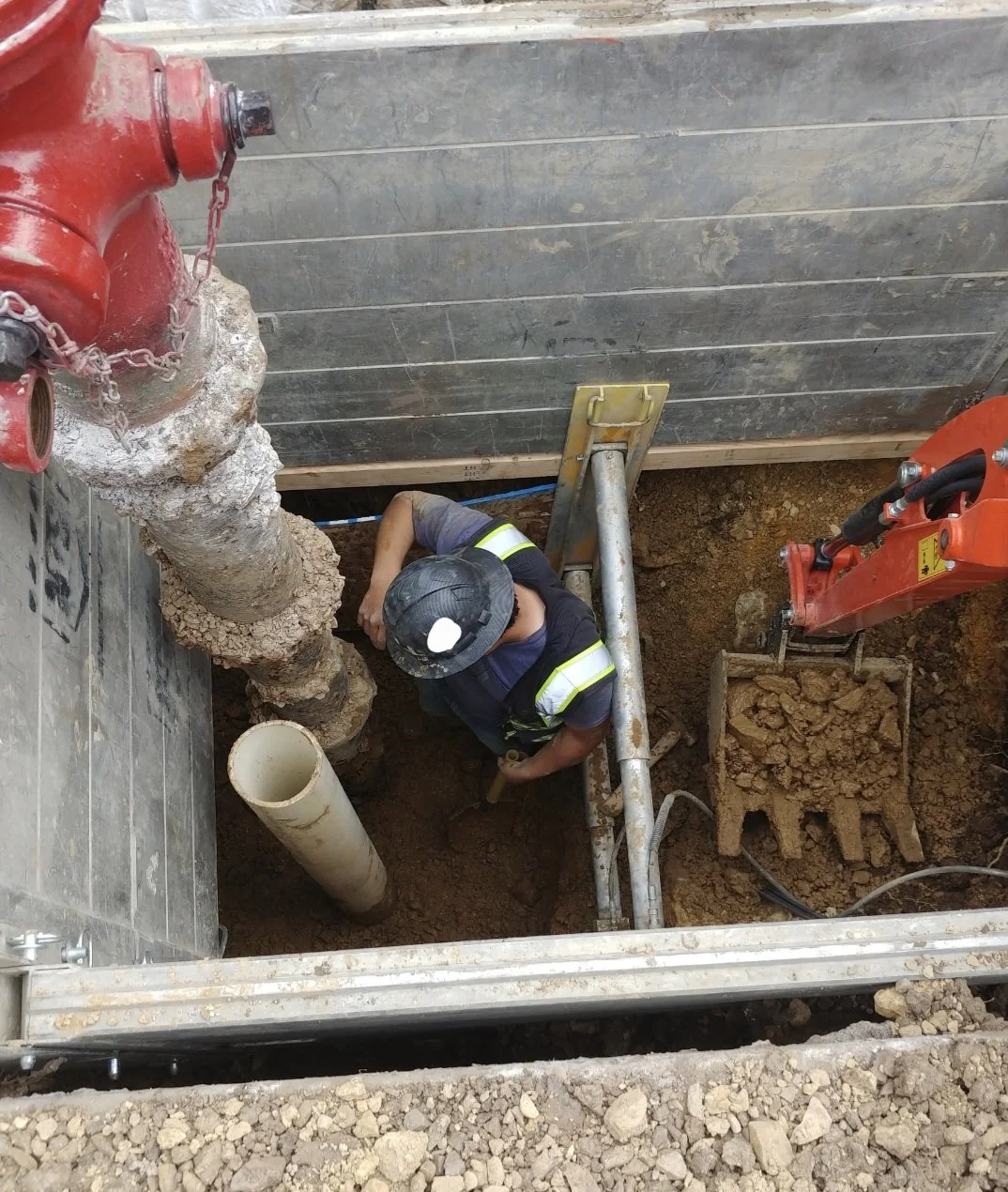 A construction worker wearing a safety helmet and vest working in an excavated trench with shoring to install or repair fire hydrant, with construction equipment and dirt around him.