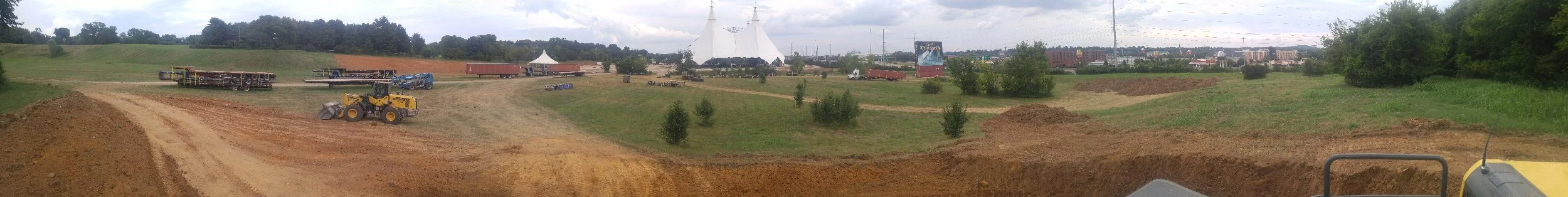 A panoramic view of a construction site with dirt mounds, construction machinery, trucks, and a large white tent in the background.