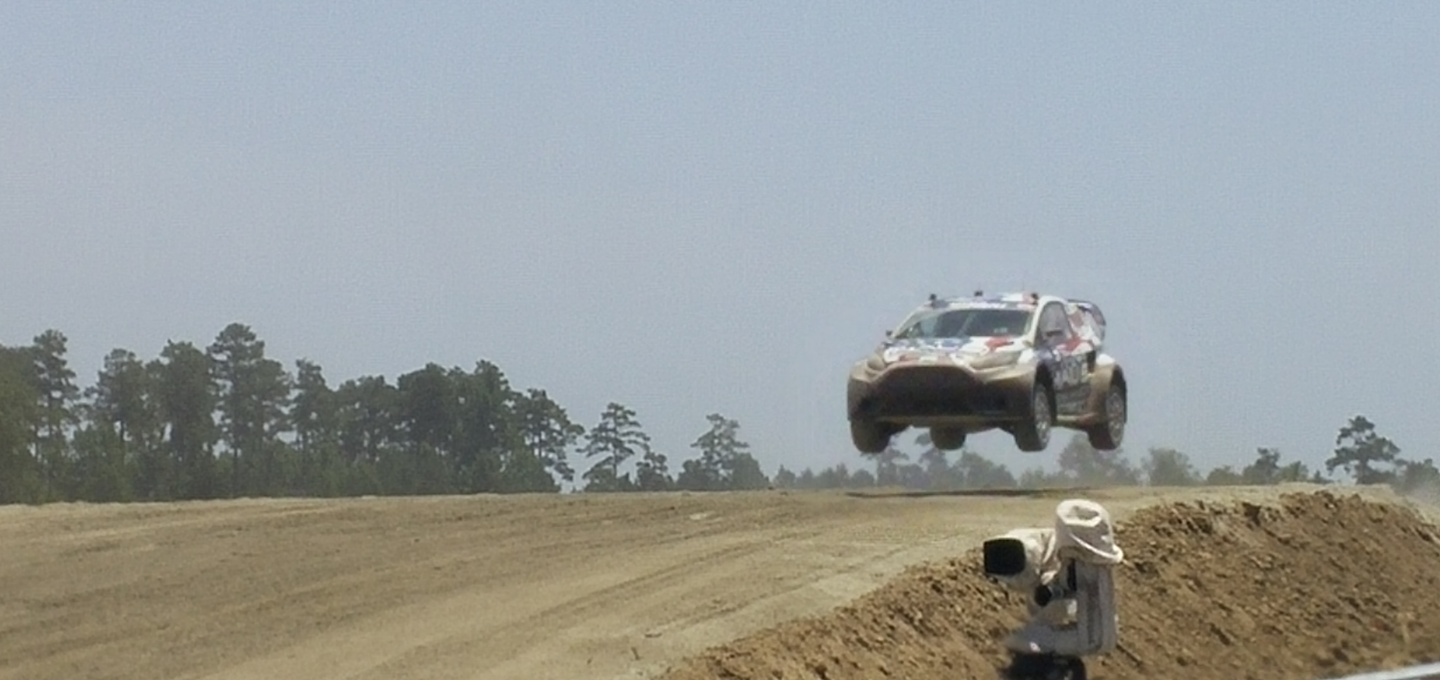 A rally car airborne over a dirt track made by Bendy Rock with dust and trees in the background.